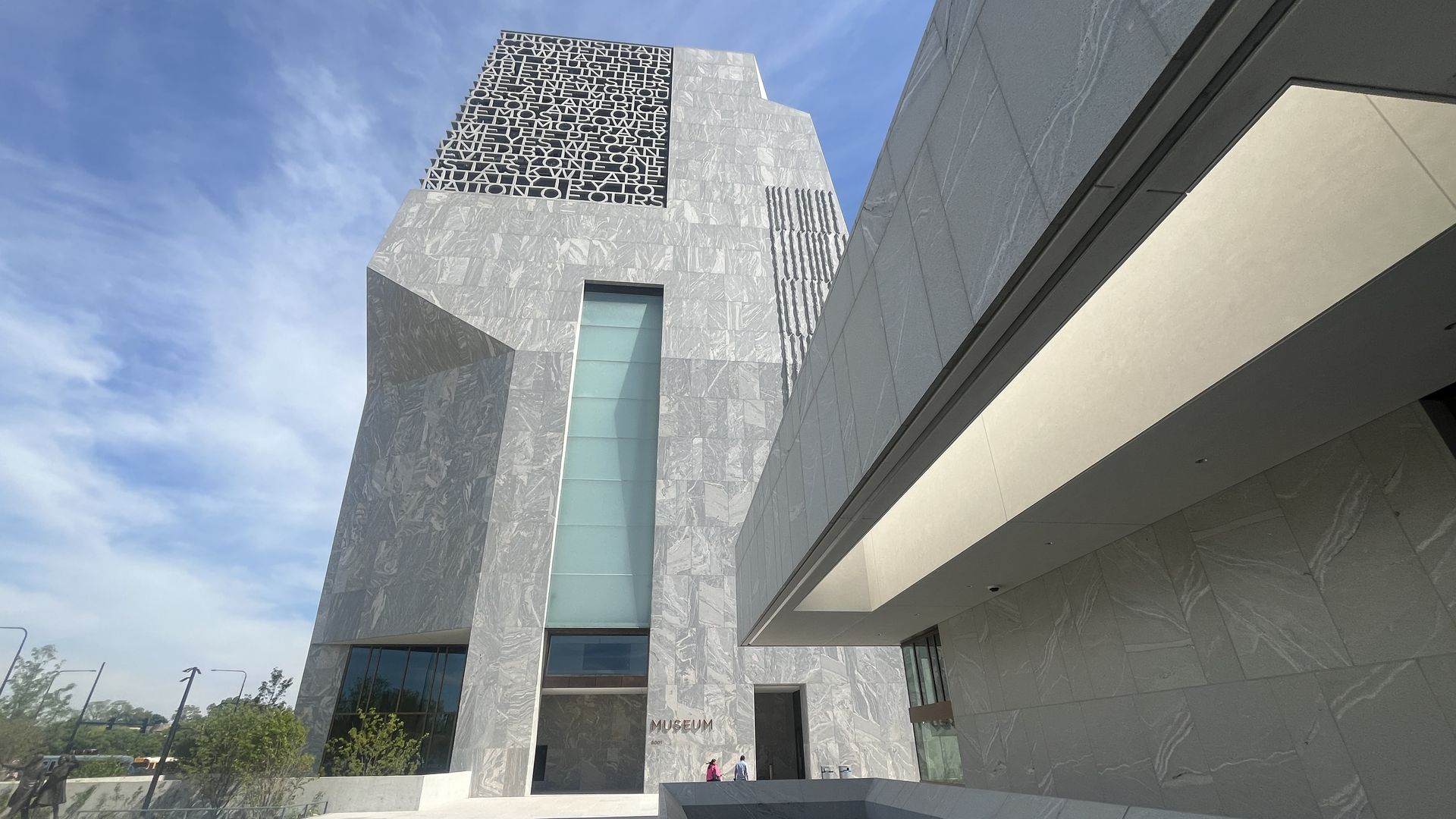 Modern silver marble museum facade with angular blocks and a large black square panel of letters. Glass windows, a covered entrance, and a few people near the doorway under a blue sky.