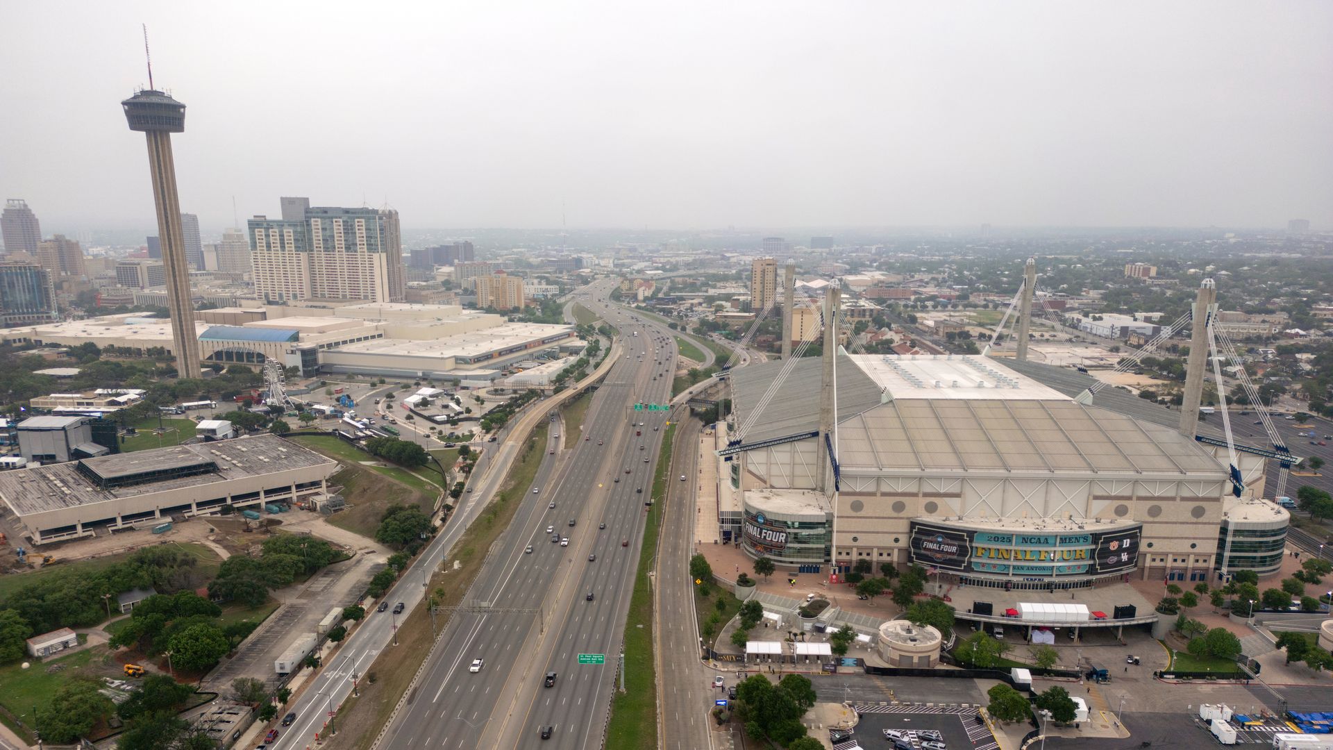 The San Antonio skyline, including the Alamodome and the Tower of the Americas, is seen against a gray, cloudy sky. Interstate 35 runs through the middle of the photo.