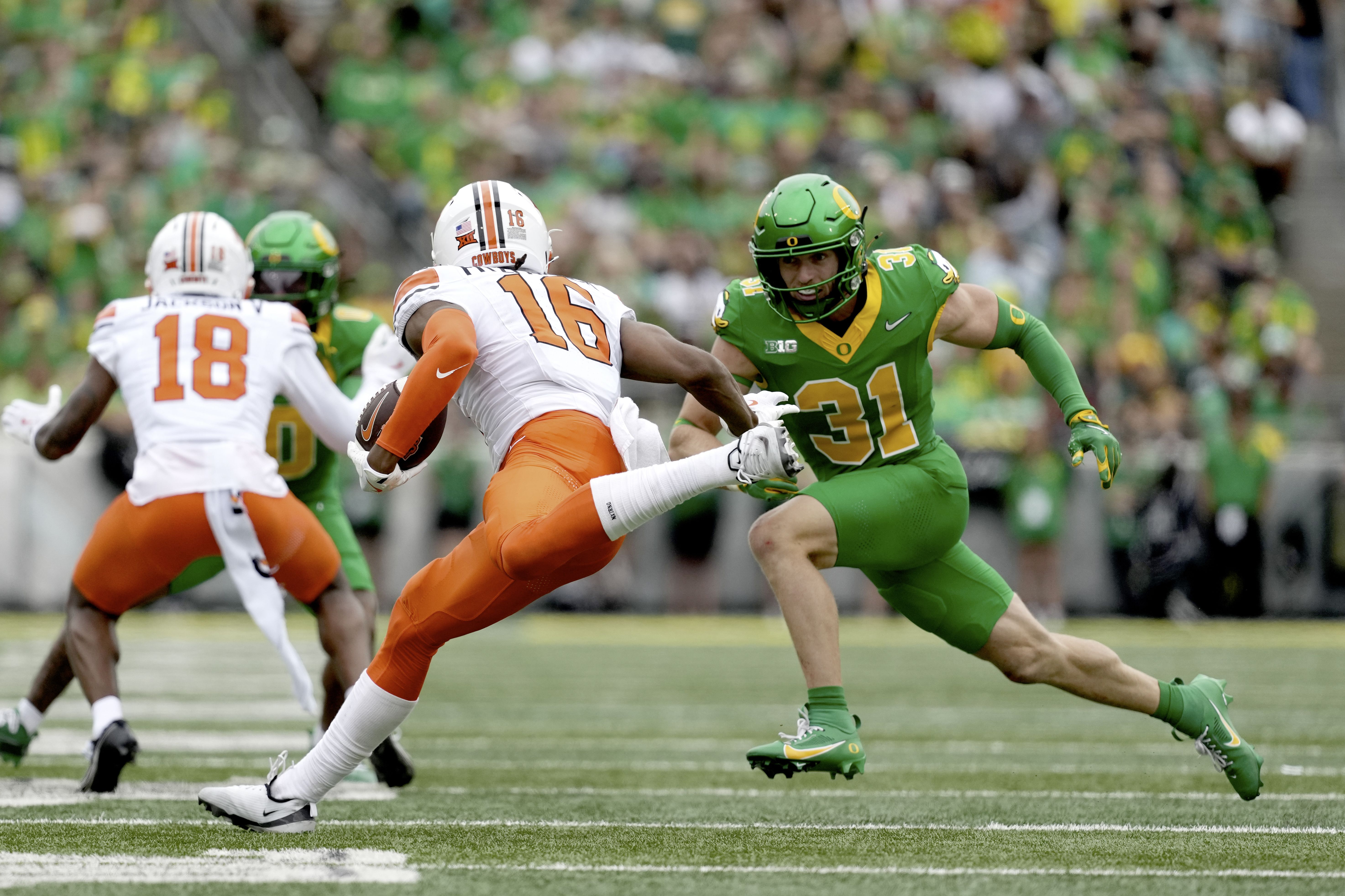 College football action: a Cowboys defender in white and orange (#16) rushes toward a Ducks defender in green (#31) as a packed, green-clad stadium watches.