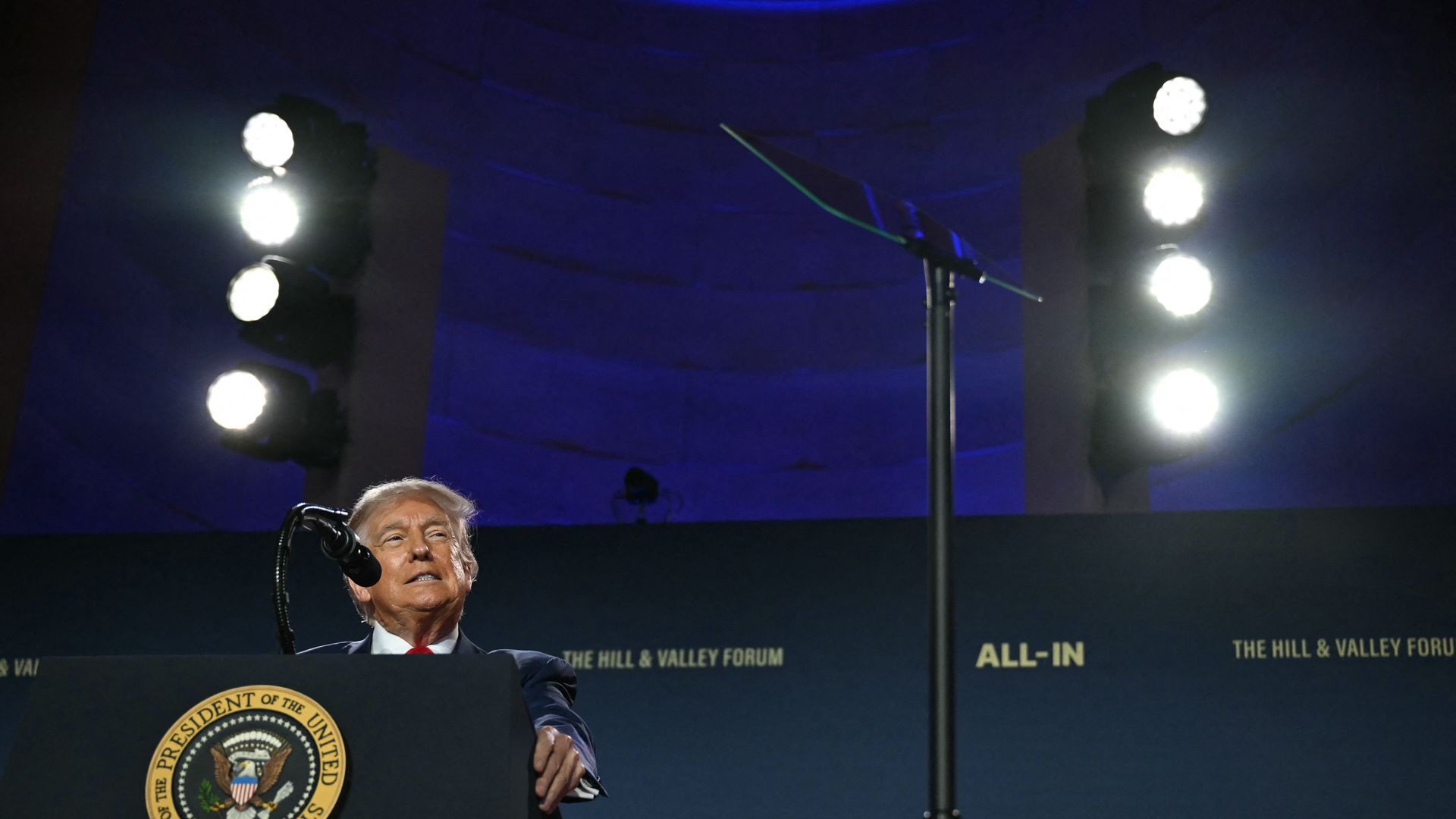 Donald Trump speaks from a podium displaying the Seal of the President of the United States against a dark blue backdrop reading "The Hill & Valley Forum" and "ALL-IN" under bright stage lights.