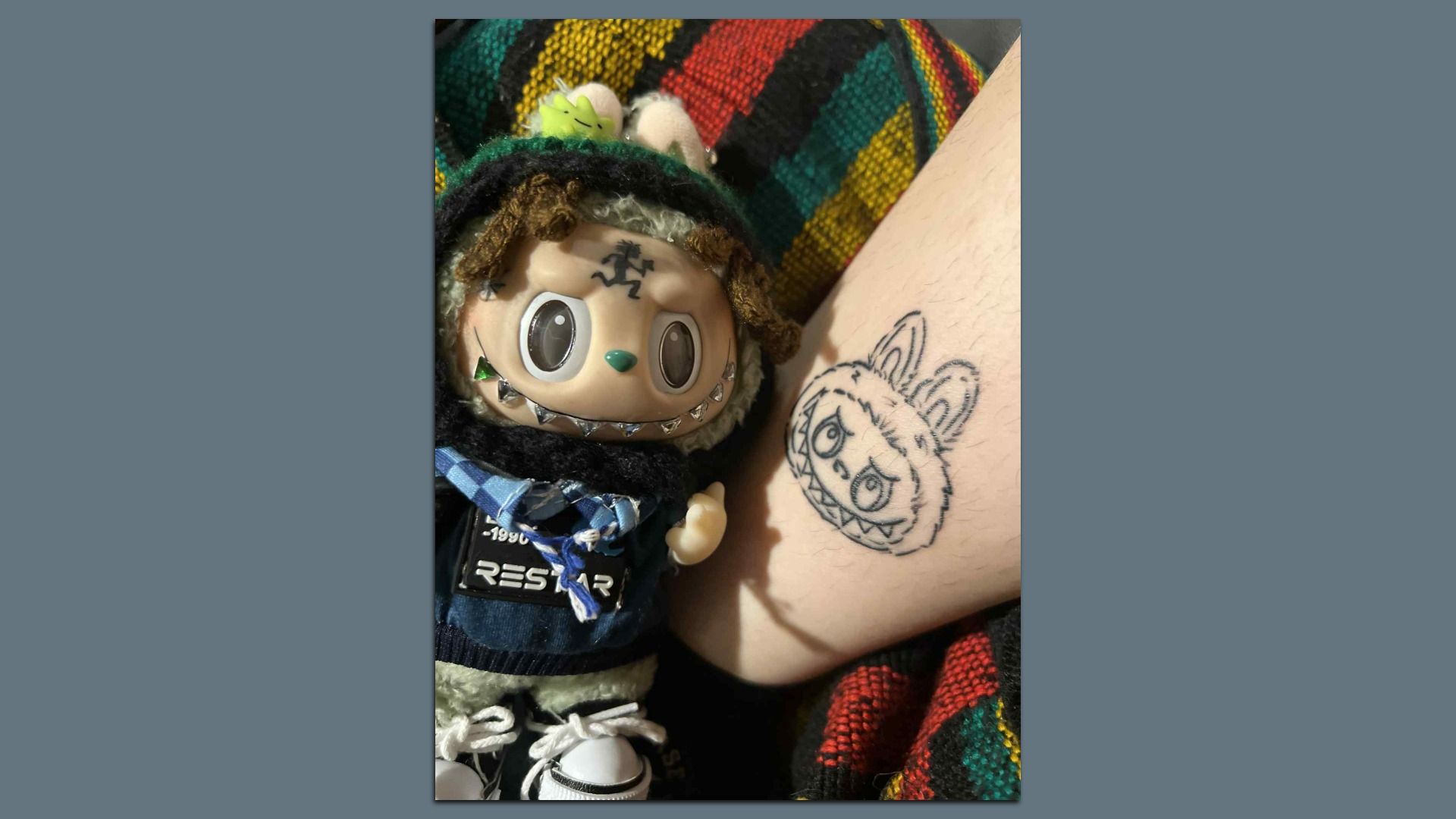 A Labubu doll sits next to a person's leg with a tattoo of the Labubu doll's face on it.