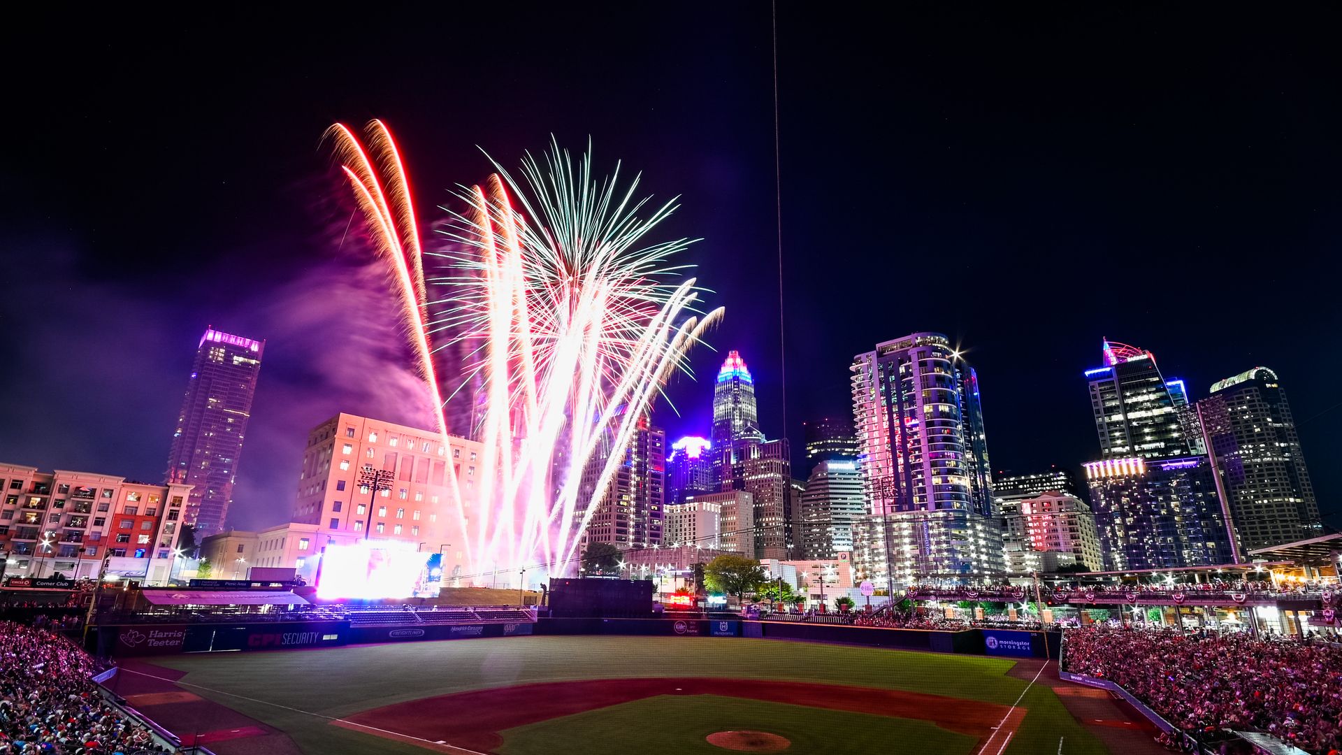 Fireworks illuminate the night sky above a baseball stadium packed with spectators, with a brightly lit city skyline in the background.