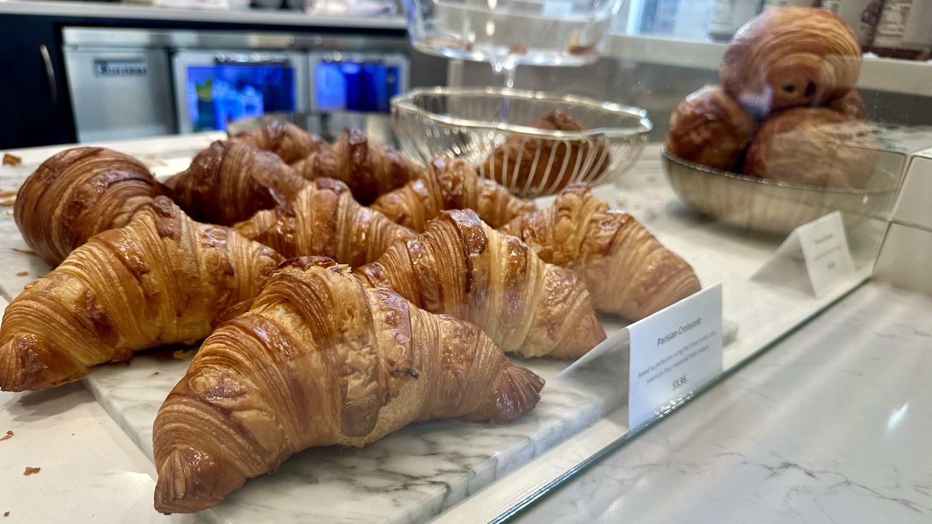 Rows of croissants on a white stone counter behind clear glass, with a pile of pains au chocolat in the  right background.