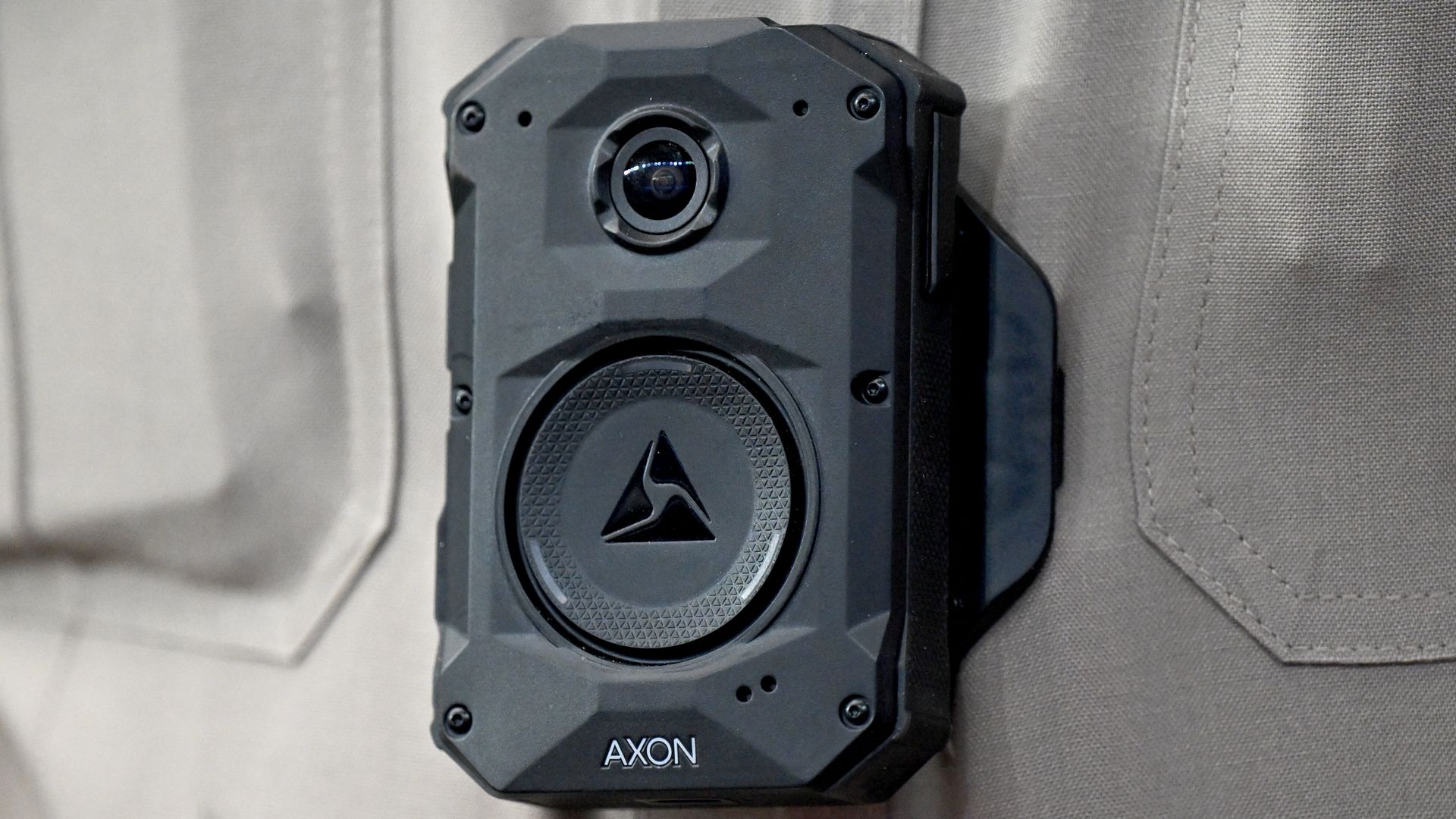 Rugged black Axon body camera with a circular lens near the top and a textured circular emblem featuring a triangular logo; mounted on gray fabric.
