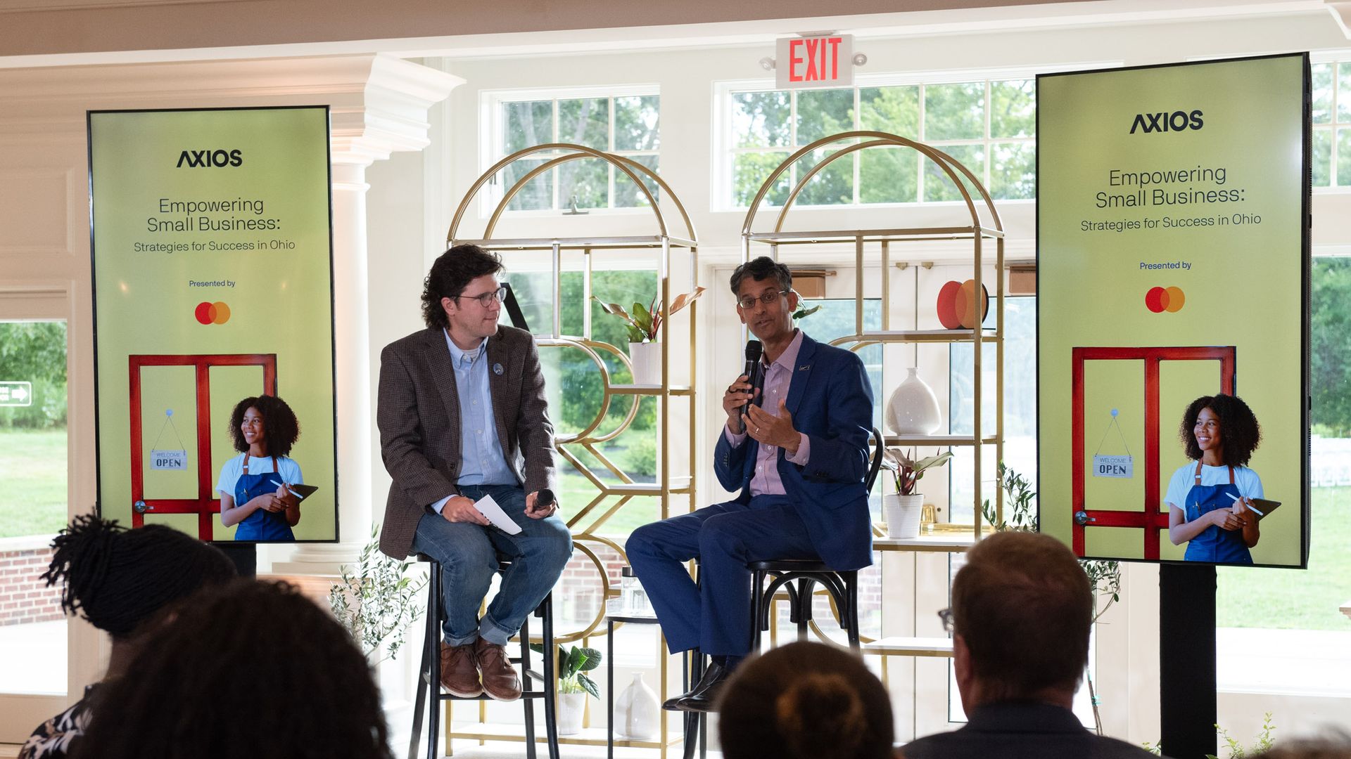 Two men on stools speaking at an indoor event about empowering small business in Ohio, with audience in foreground and two screens displaying event info and a woman in blue apron outside.