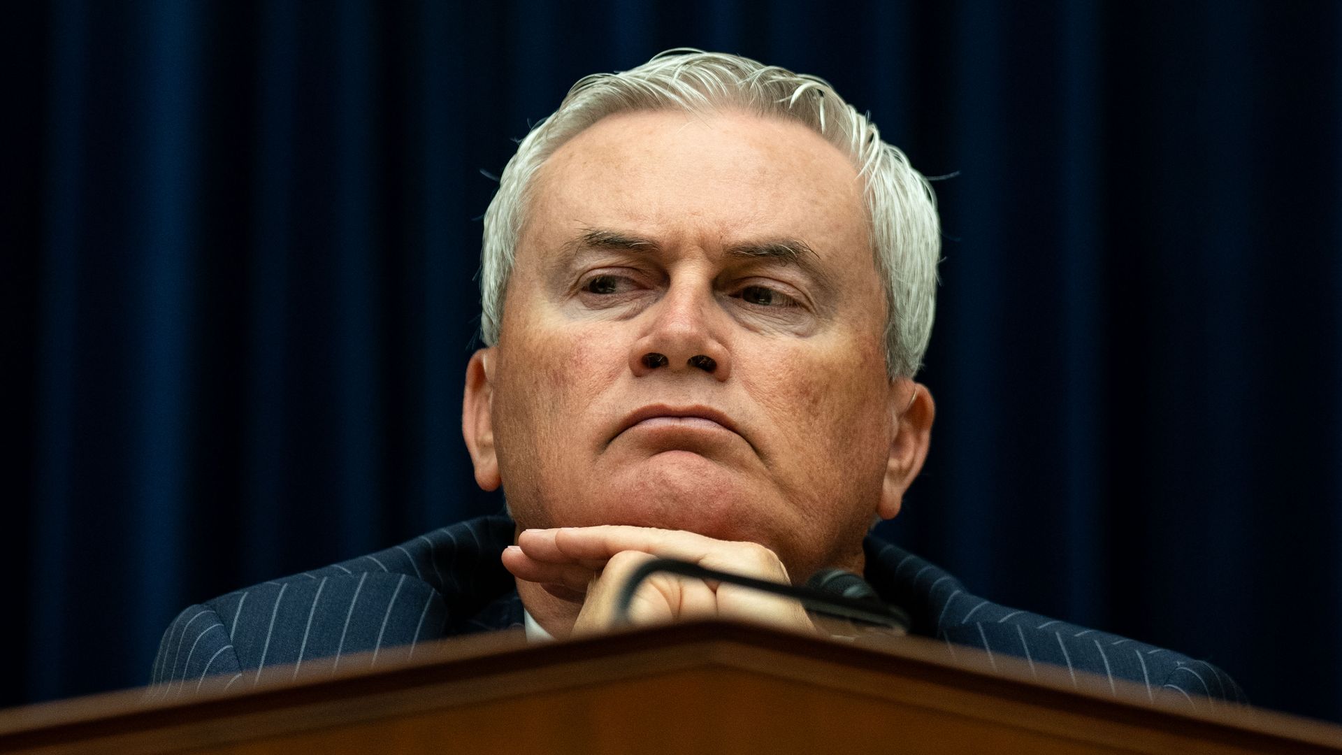 James Comer, wearing a pinstripe suit with his head on his hands, sitting at a wooden dais in front of a blue curtain.