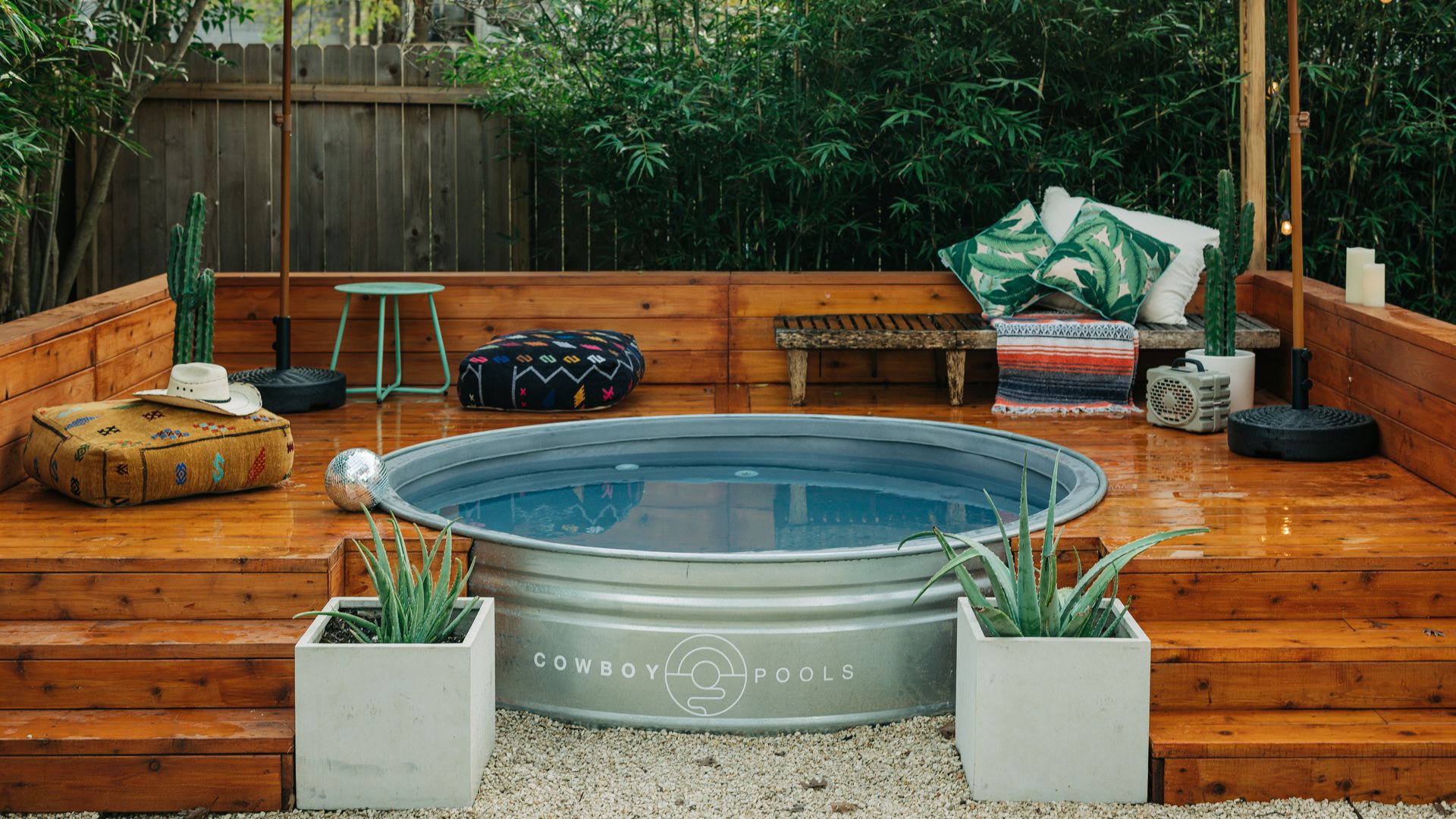A circular metal pool filled with water with a Cowboy Pools logo on it surrounded by a wooden deck flanked by two square-shaped pots with spikey succulents inside. On the porch is a bench, table, cushion with a cowboy hat on top, cactus and two umbrellas. Behind it is a wooden fence and trees.