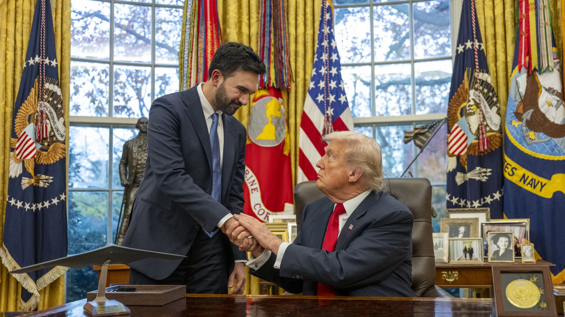 Donald Trump, sitting at the resolute desk, shakes hands with Zohran Mamdani, who is standing beside him in the Oval Office.
