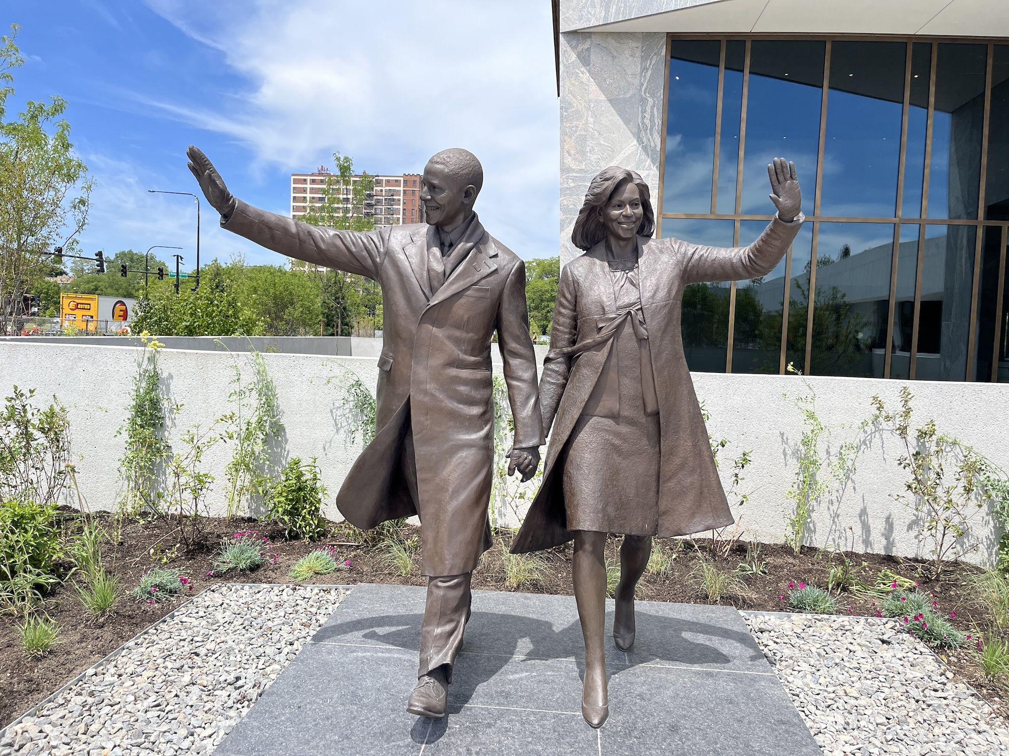 Bronze statues of Barack and Michelle Obama walking hand in hand; Barack waves with his outstretched arm, Michelle raises her hand. A modern glass-front building and blue sky behind.