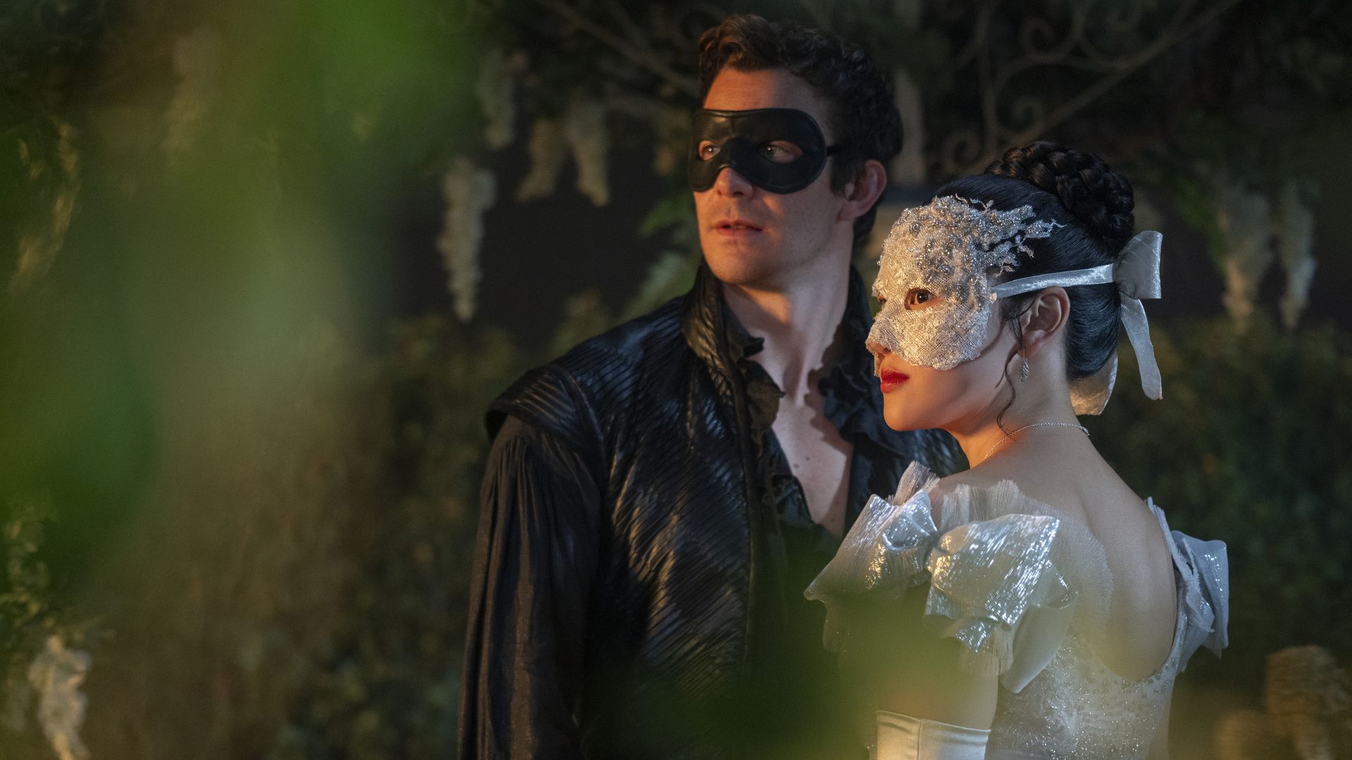 A man in a black leather costume and mask stands beside a woman in a white dress with puffy sleeves and a decorative white mask, both looking to their left in a dim garden setting.