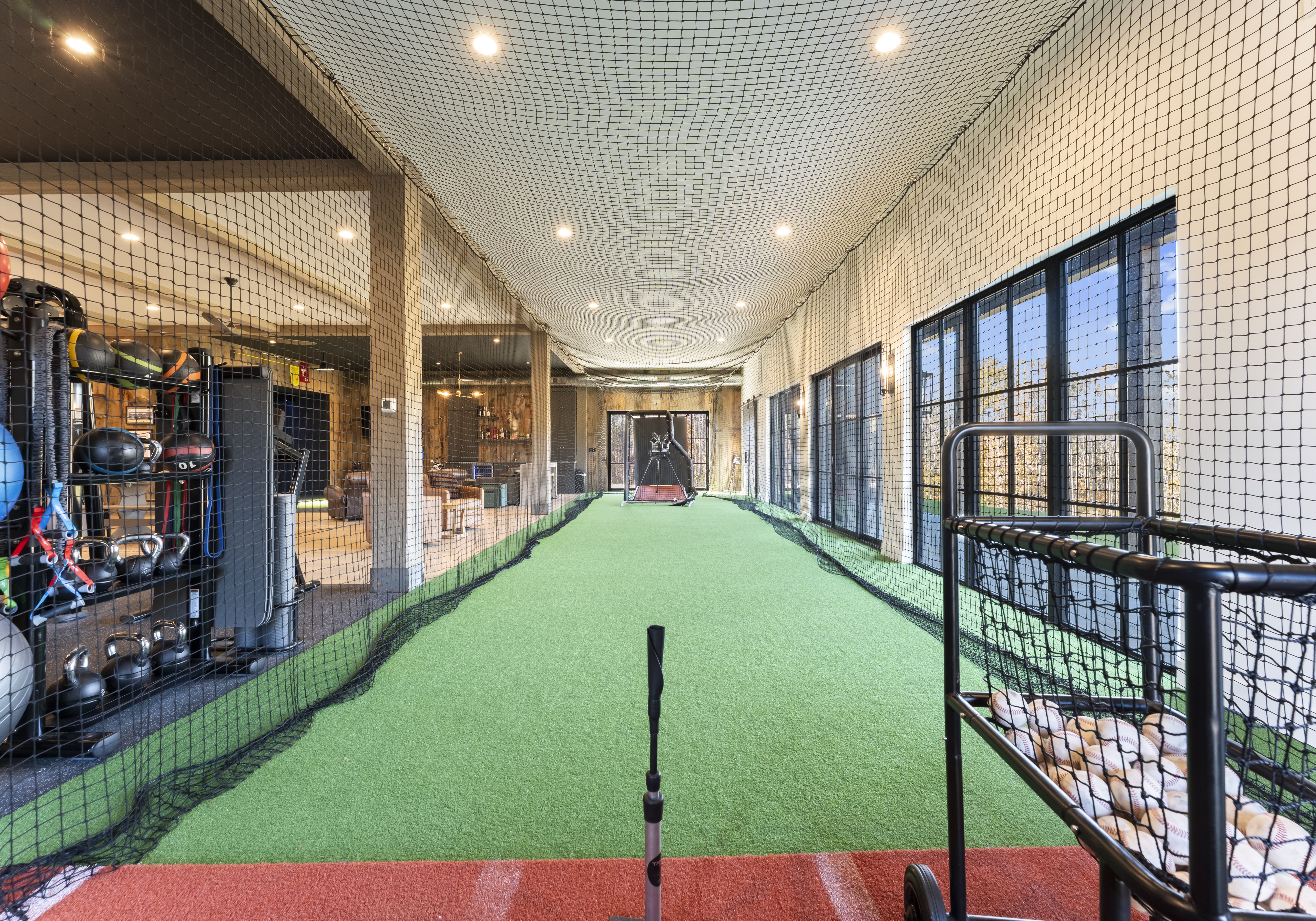 Indoor athletic training space with a long green turf lane enclosed by netting. A baseball bat stands upright in the foreground; gym equipment on the left, large windows on the right, and a pitching/batting area at the far end.