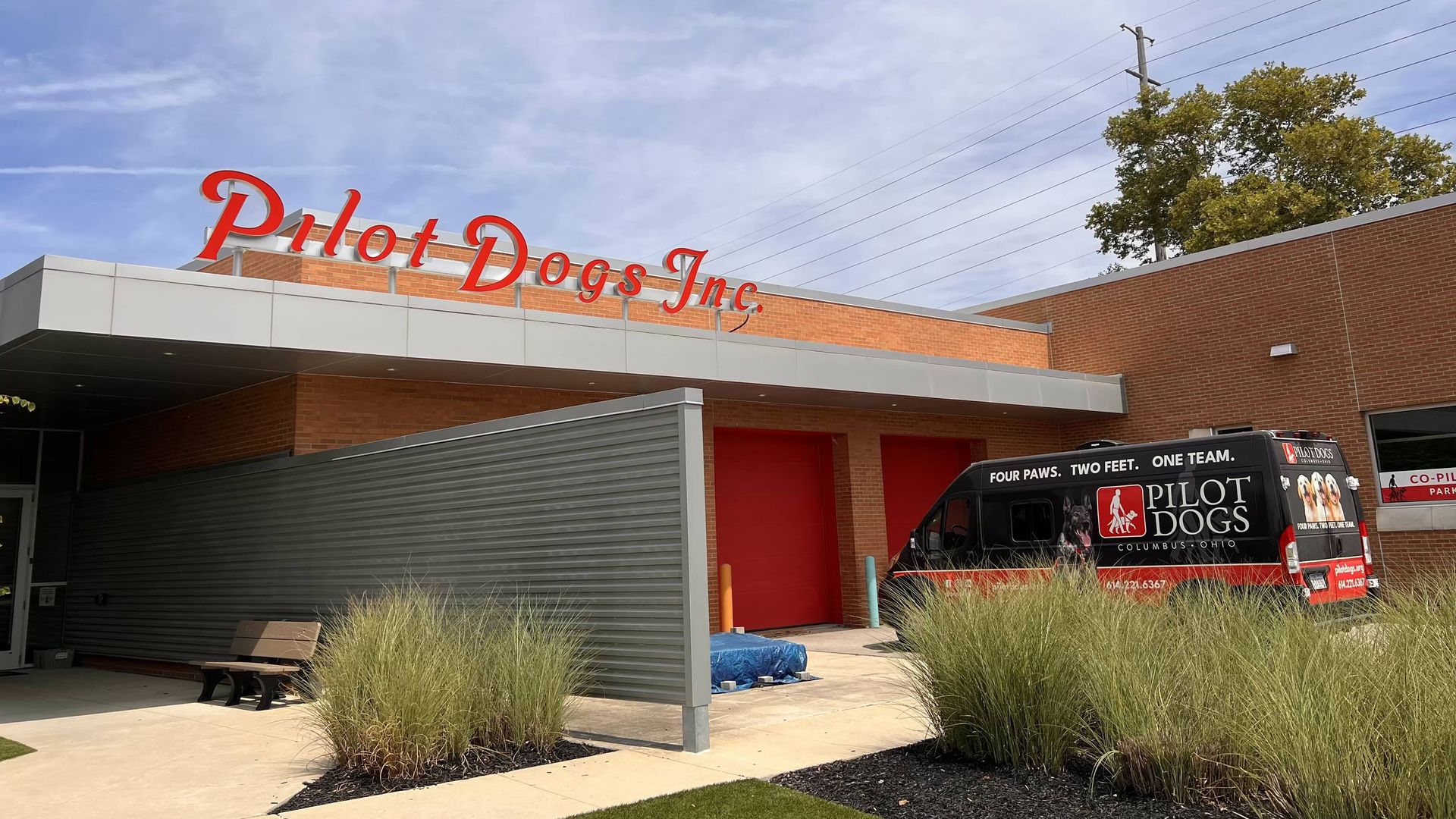 Building with red sign reading Pilot Dogs Inc. and van with Pilot Dogs logo and slogan, surrounded by grass and a partly cloudy sky in the background.