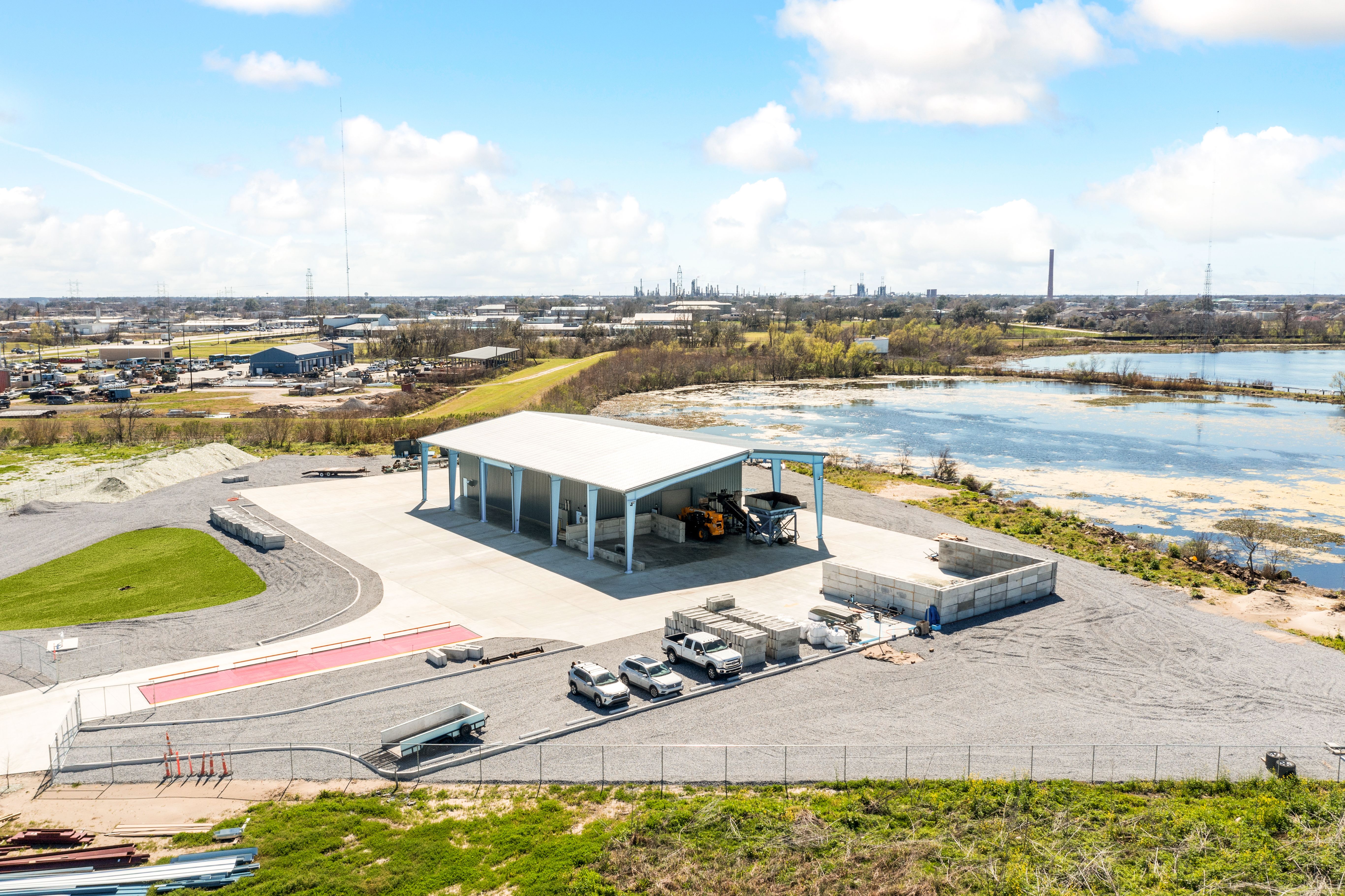 Wide aerial view of an industrial park: blue warehouse, yard with vehicles and stacked materials, a pond on the right, distant refinery with a tall smokestack, under a bright blue sky.
