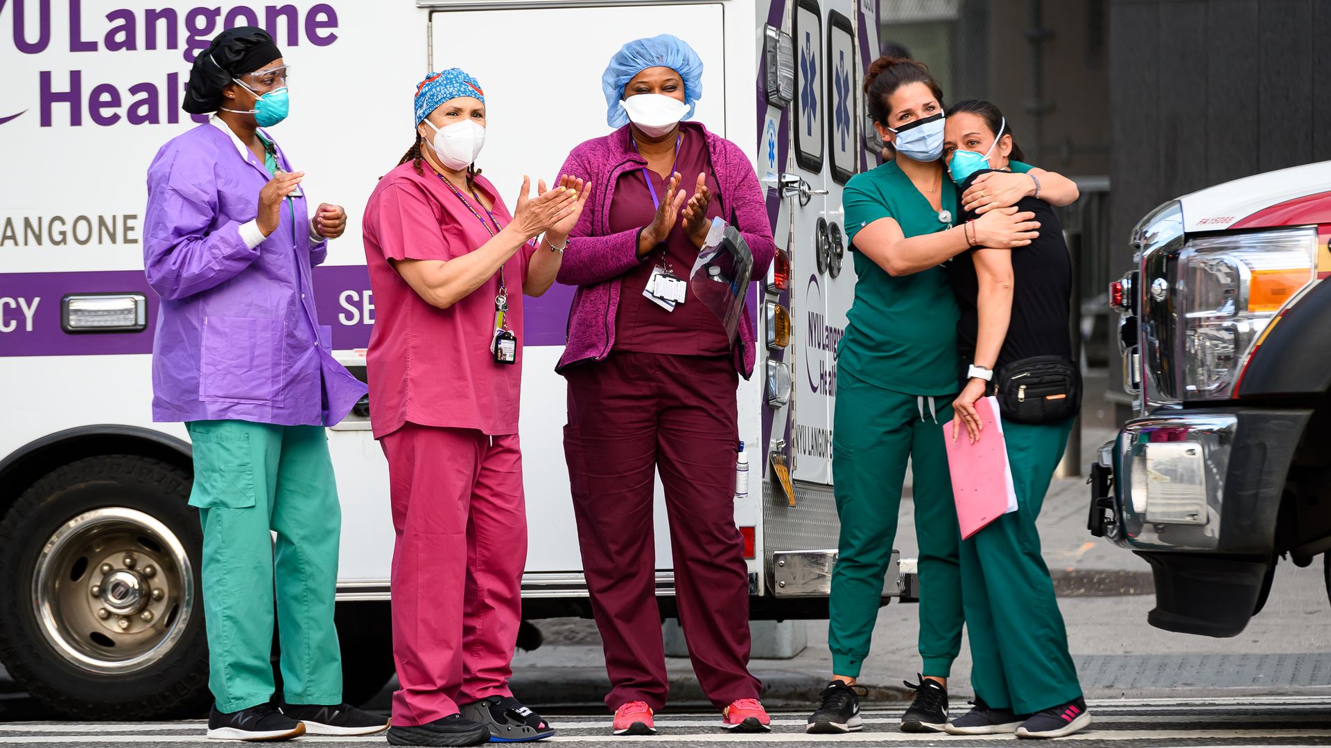 Health care workers stand in front of a hospital wearing face masks and scrubs as they clap