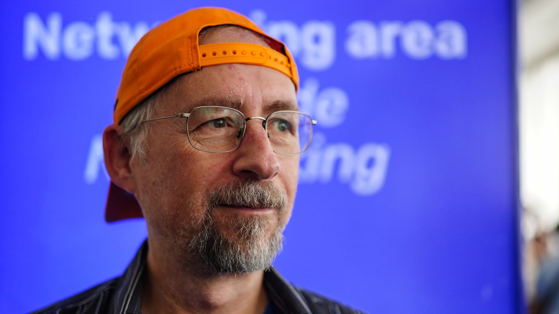 Portrait of a middle-aged man with gray beard and glasses, wearing an orange baseball cap backward and a blue shirt. He stands before a bright blue backdrop with blurred white text.