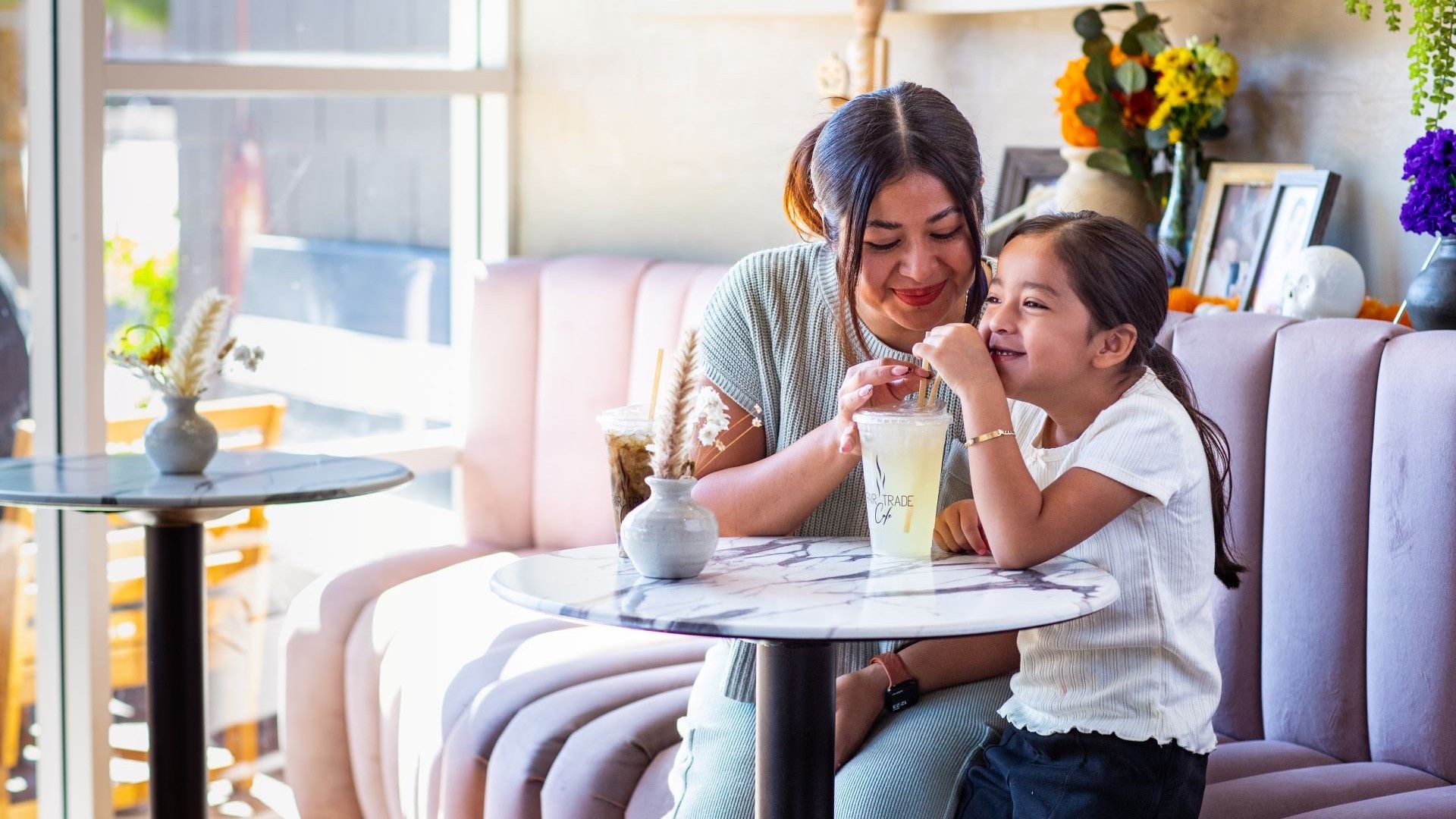 Two women, a mother and daughter, sit at a marble cafe table in a pastel pink booth, sharing a light drink with straws as sunlight streams in; bright flowers and frames decorate the cozy background.