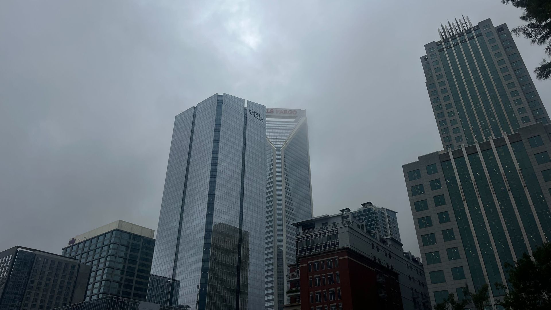 City skyline with tall modern glass and concrete buildings under overcast, cloudy sky, creating a gray, moody atmosphere.