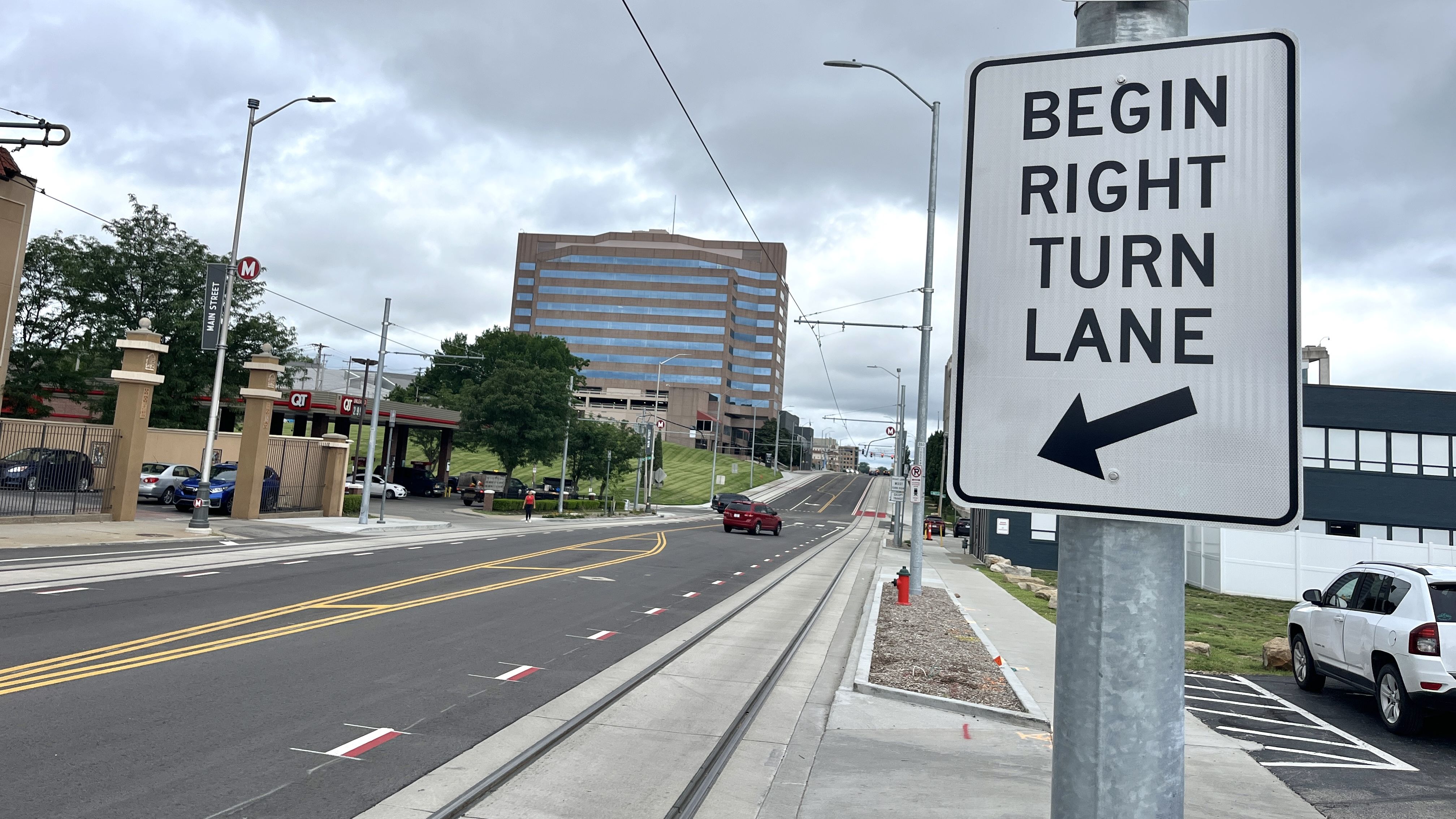 Street scene with a large white sign reading "BEGIN RIGHT TURN LANE" and a black arrow pointing left, a building with blue windows in the background, cars parked and driving on a cloudy day.