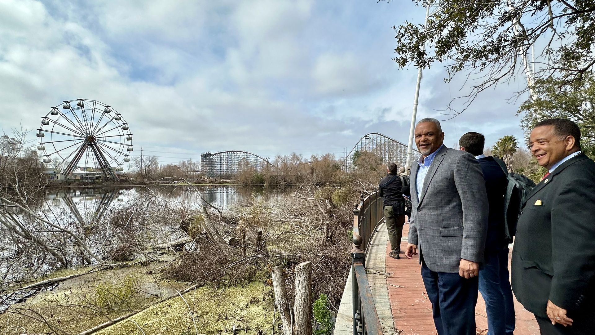Group of men in suits stand along a boardwalk beside a marshy waterway, with a large Ferris wheel and a roller coaster in the distance; cloudy sky and leafless trees frame the scene.