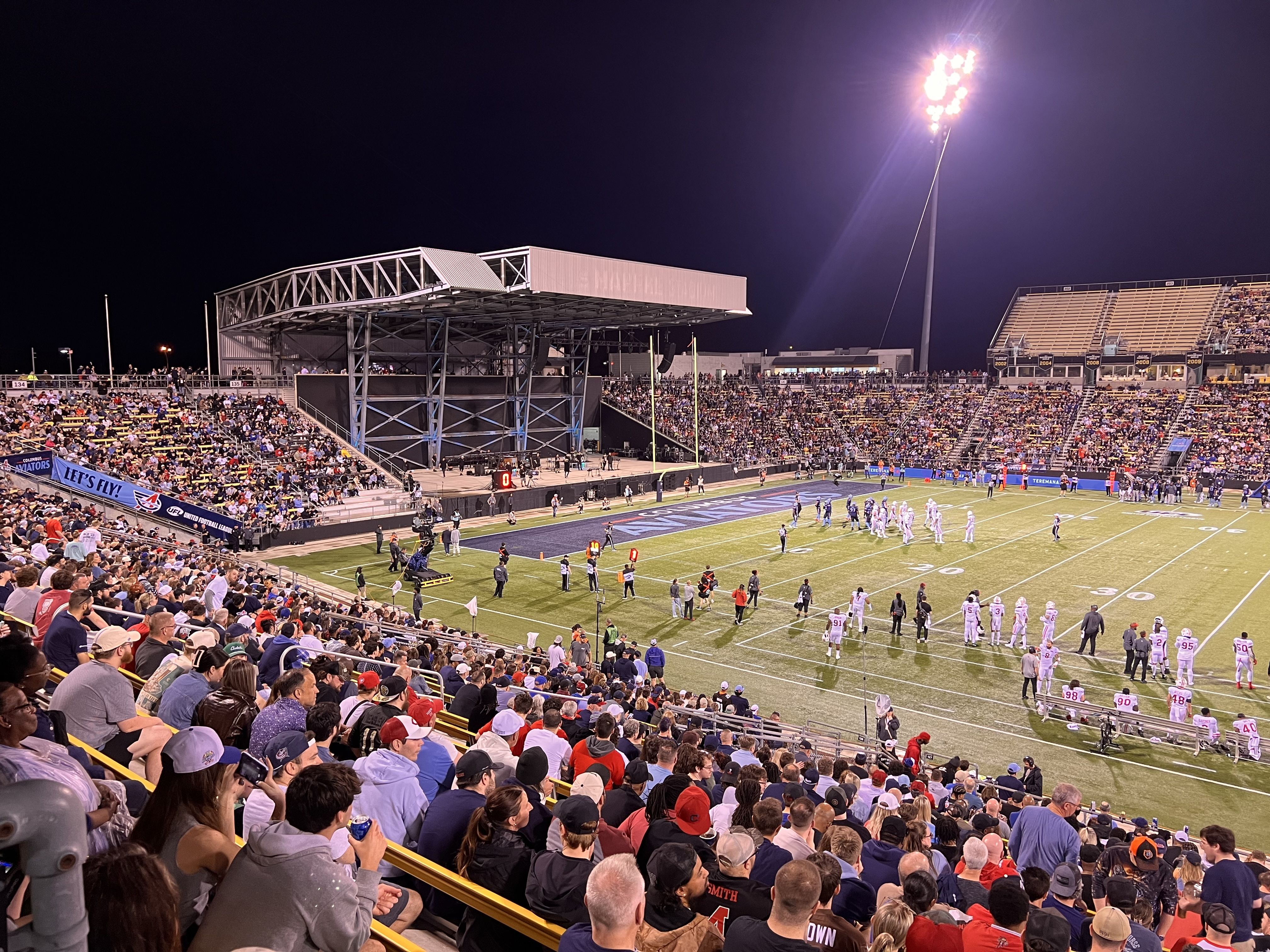 Nighttime football game in a large stadium with towering floodlights. Players in white uniforms on a green field, while a full crowd fills the stands on both sides under bright stadium lights.