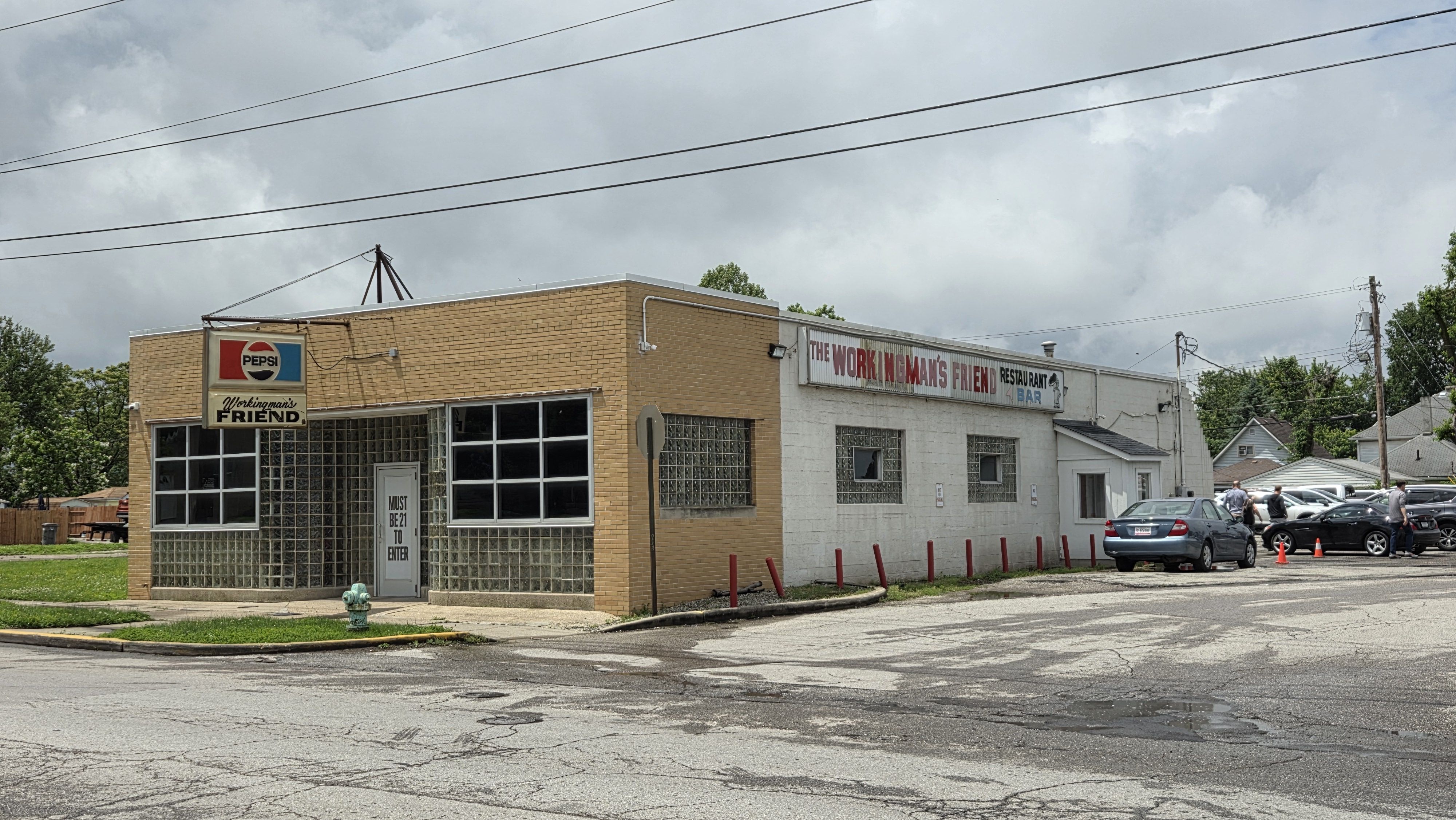 Beige brick storefront with glass block windows and a Pepsi sign reading "Workingman's Friend." A long exterior sign reads "The Working Man's Friend". Cars park nearby under power lines.