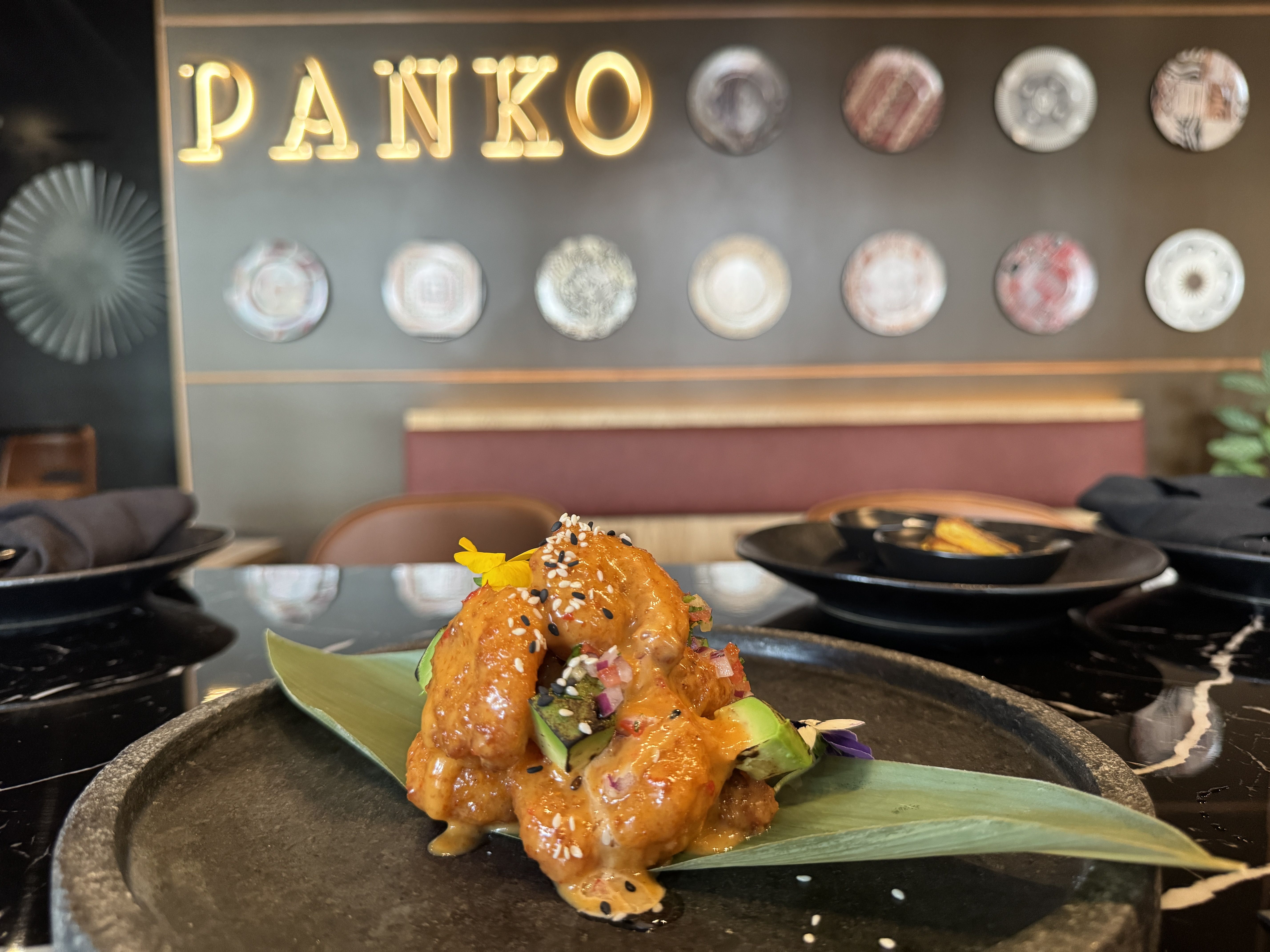 Orange-glazed shrimp with avocado chunks and sesame seeds on a green leaf on a black plate, with a restaurant interior background showing the word PANKO and decorative plates.