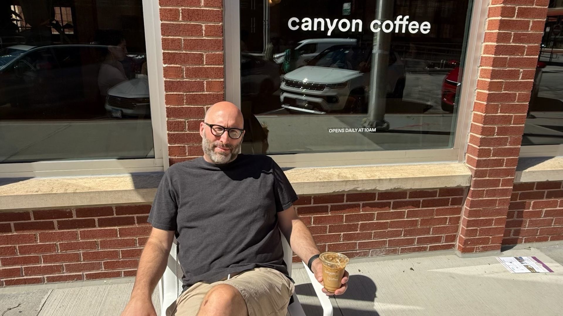 A bald man with glasses sits on a white chair outside a brick storefront. He wears a dark T-shirt and khaki shorts, holding a coffee; the window shows canyon coffee and car reflections.