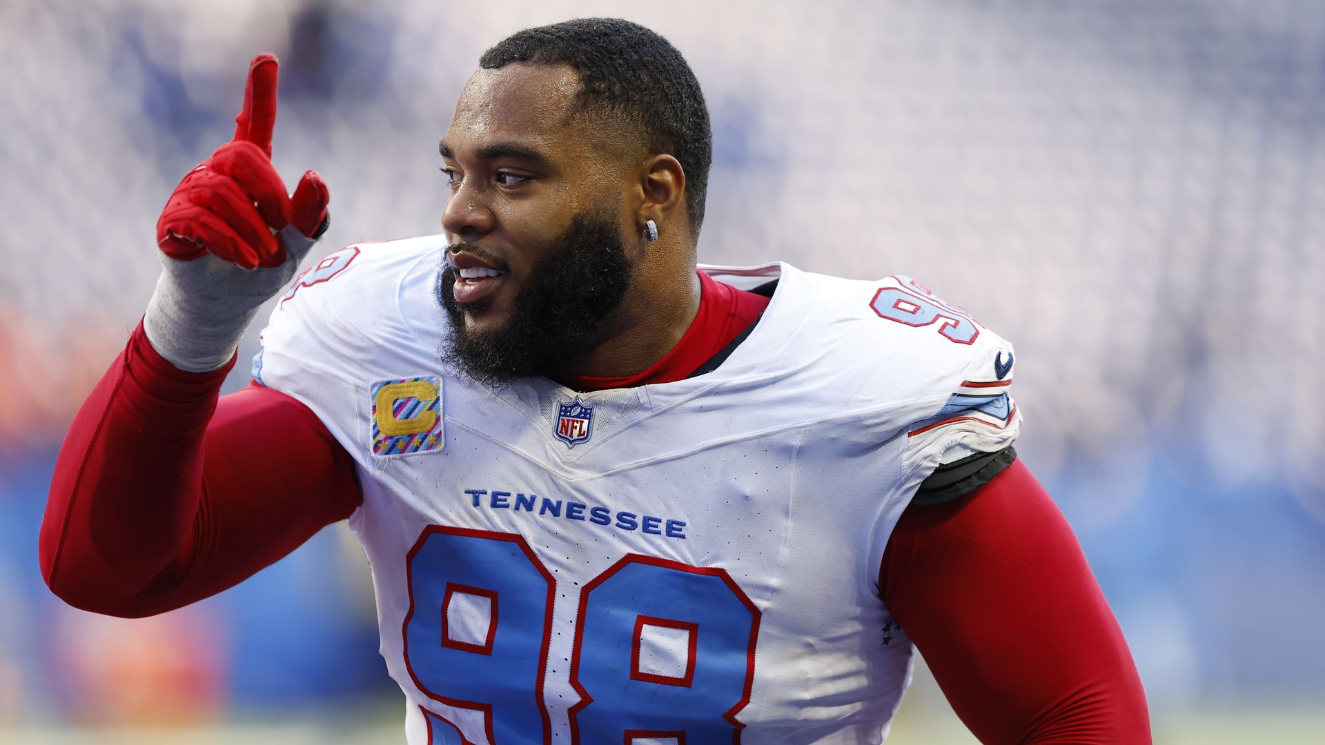 Close-up of a Tennessee Titans football player in a white jersey with blue numbers, red sleeves and gloves, pointing upward with his index finger in a stadium.