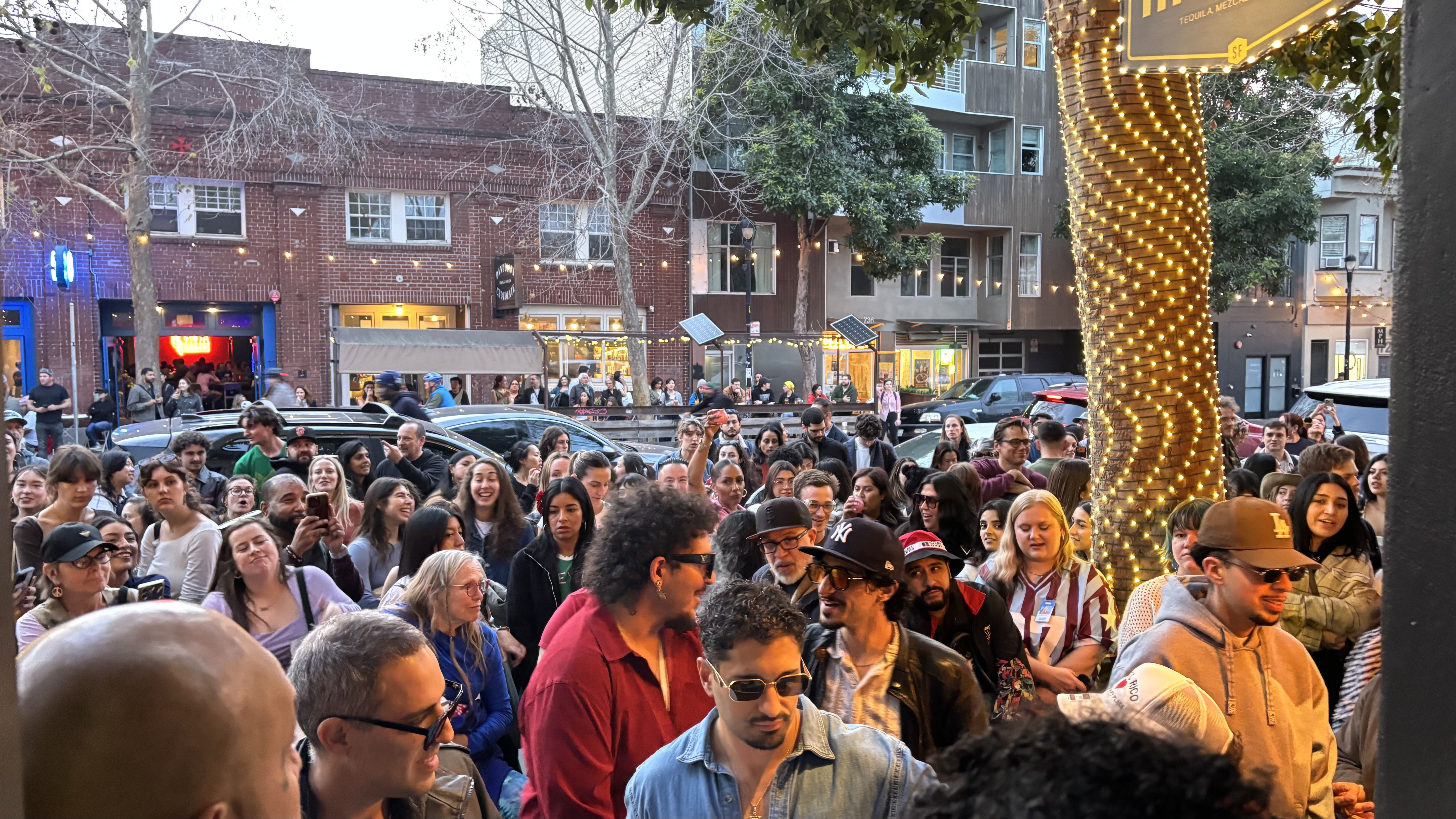 Crowd of diverse people gathered outdoors in front of buildings and cars, some wearing sunglasses and hats, tree wrapped with string lights in evening urban setting.
