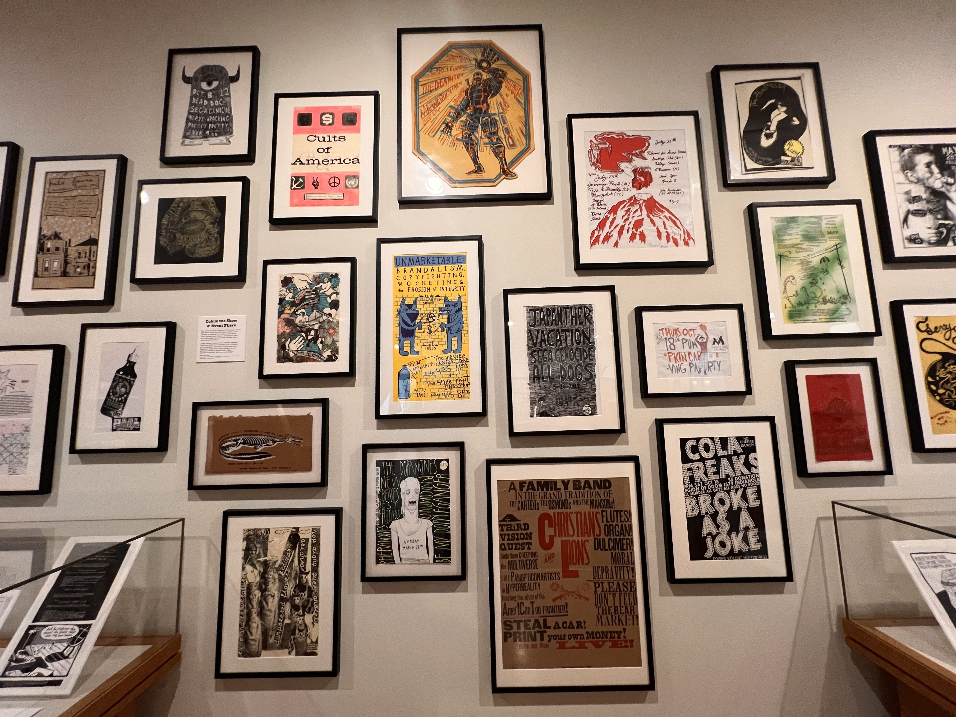 Gallery wall of black-framed posters in various sizes, arranged in a grid. Colorful prints and typography art, including a central orange hexagonal figure and a red volcano illustration.