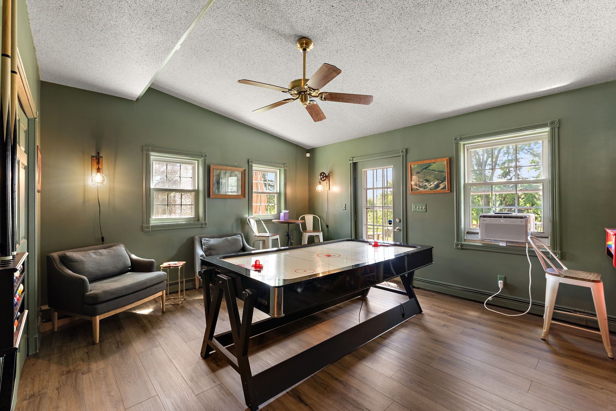 Game room with green walls, wood floor, air hockey table in center, two gray chairs with small table near windows, wall lamps, ceiling fan, and wall art with natural light coming in.