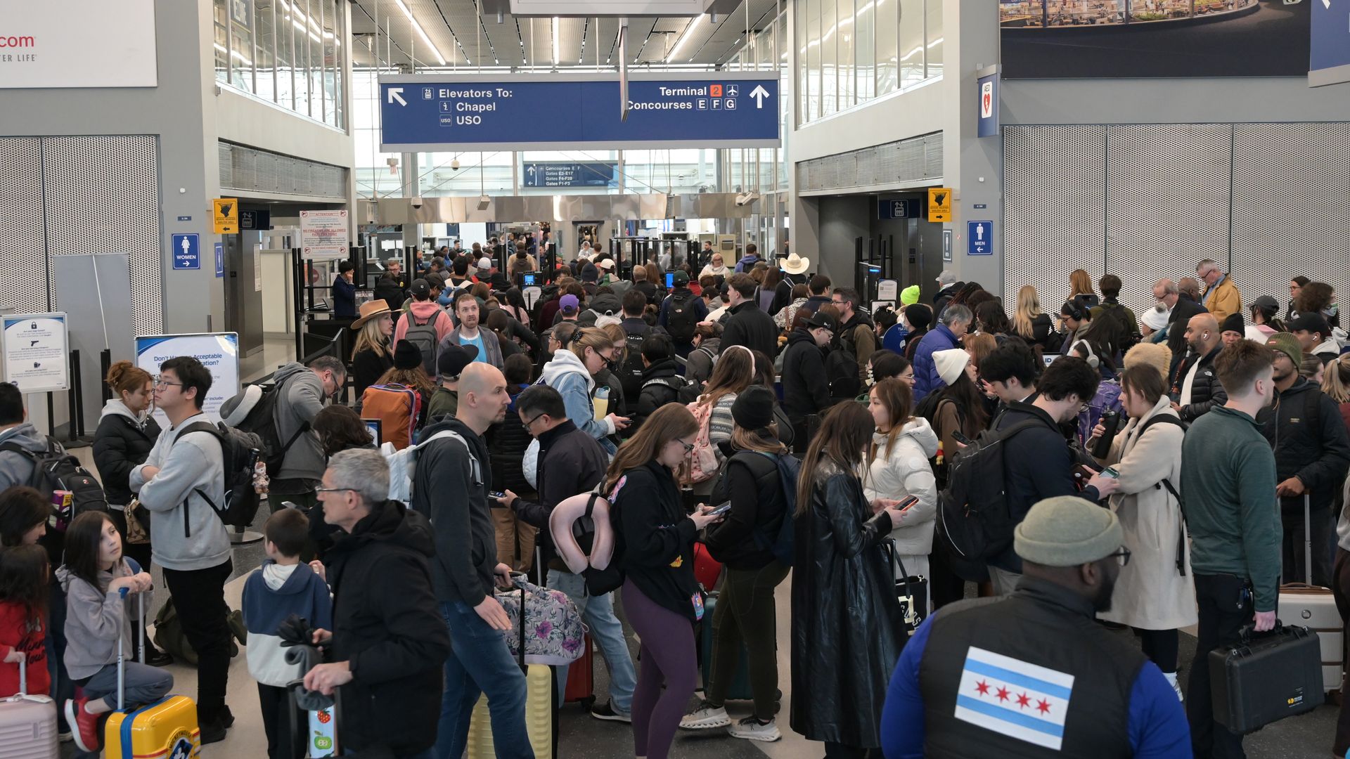 Crowded airport security checkpoint with travelers queueing and rolling luggage; blue overhead signs point to elevators, terminals, and concourses inside a bright, modern terminal.