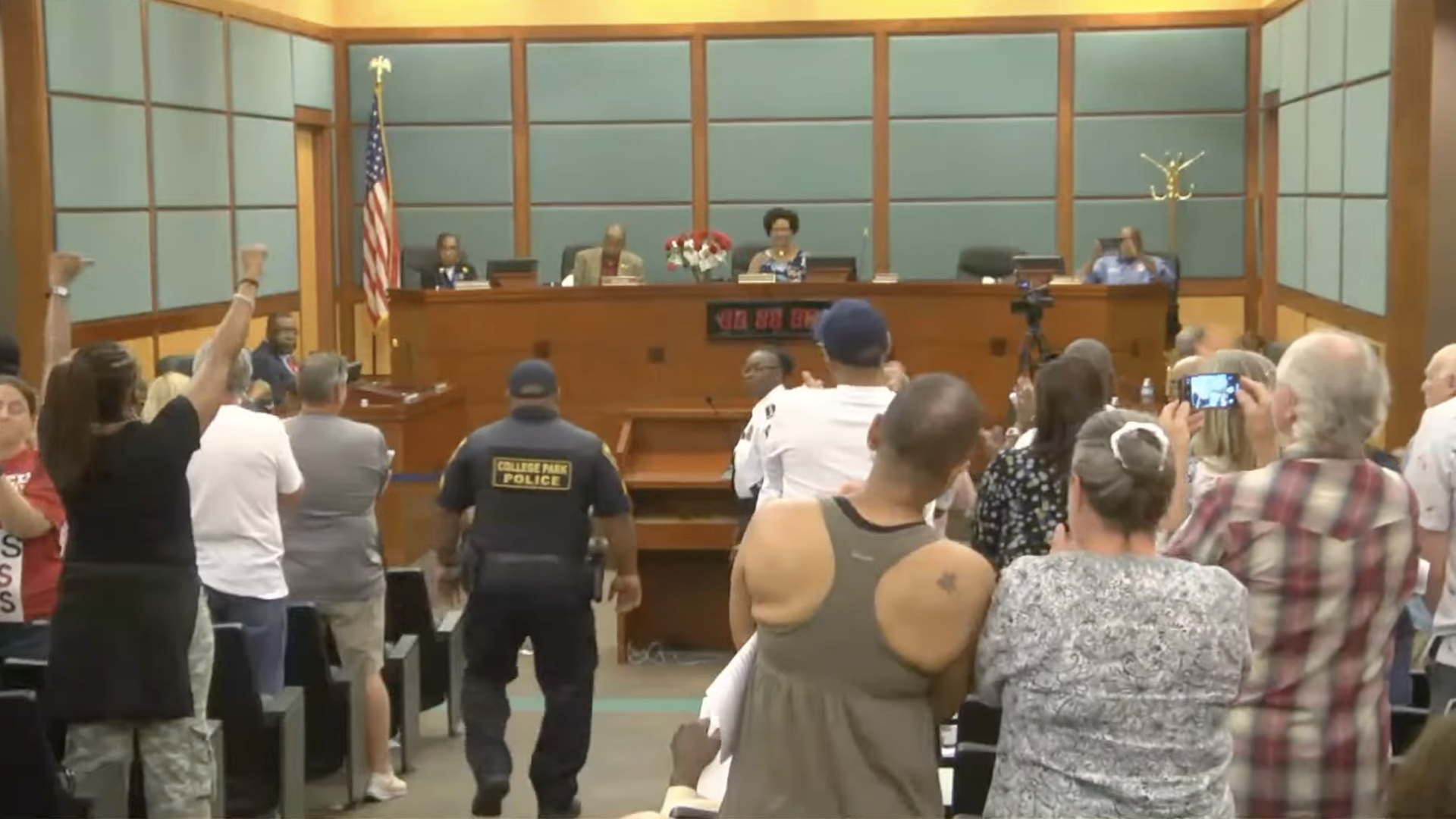 A police officer walks down the aisle of a crowded local government meeting where audience members are clapping