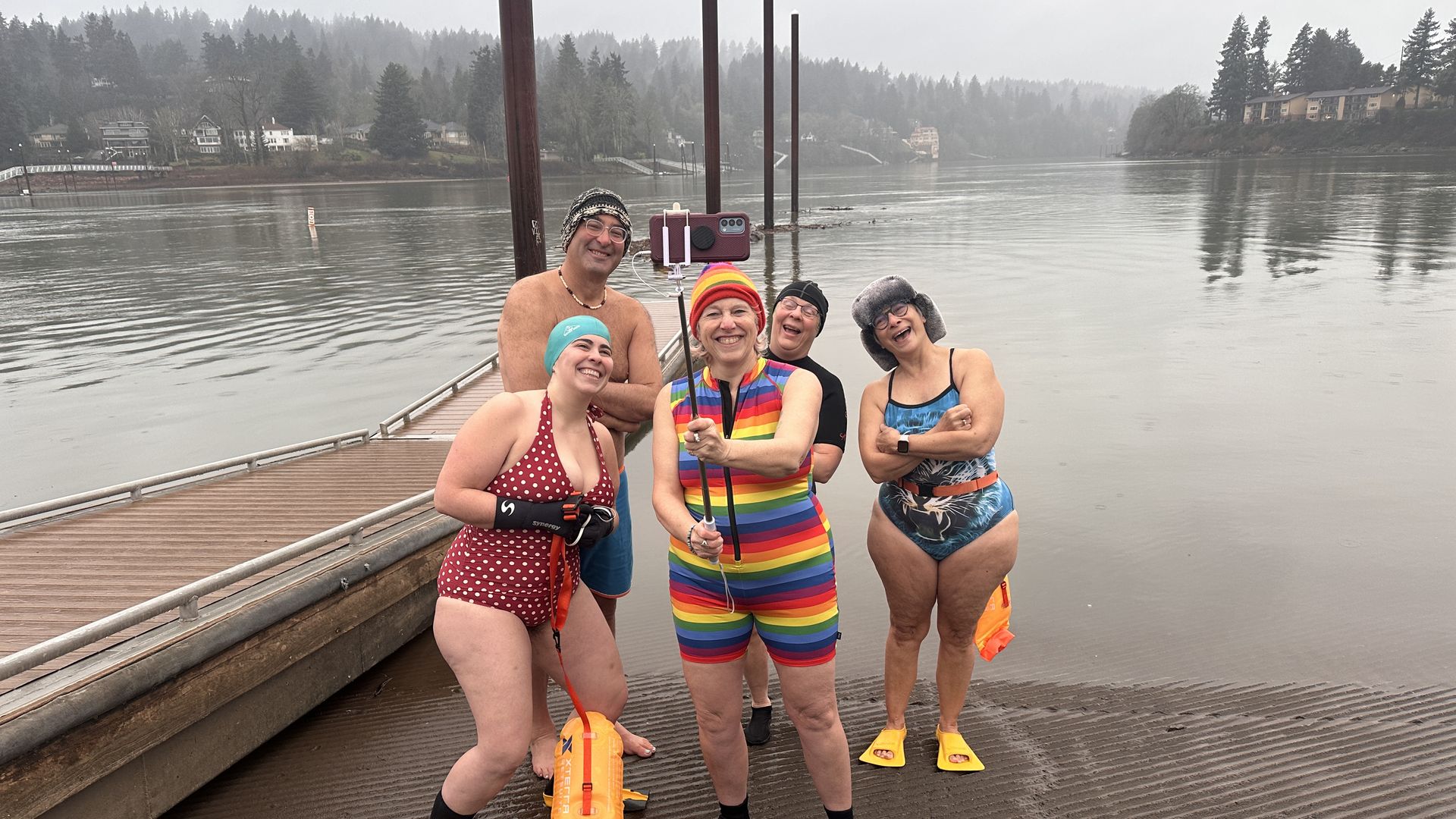 Four women and one man in bathing suits taking a selfie with a selfie stick at dawn on a boat ramp to the Willamette River in February