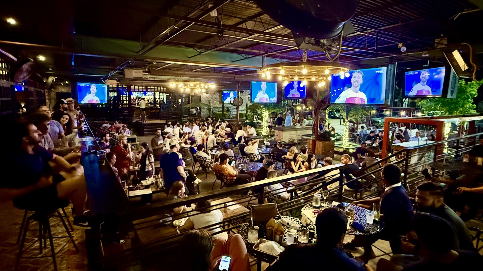 Wide interior of a lively bar/restaurant at night: many tables filled with people, multiple screens showing a man on TV, warm hanging lights, exposed ceiling, and a railing in the foreground.