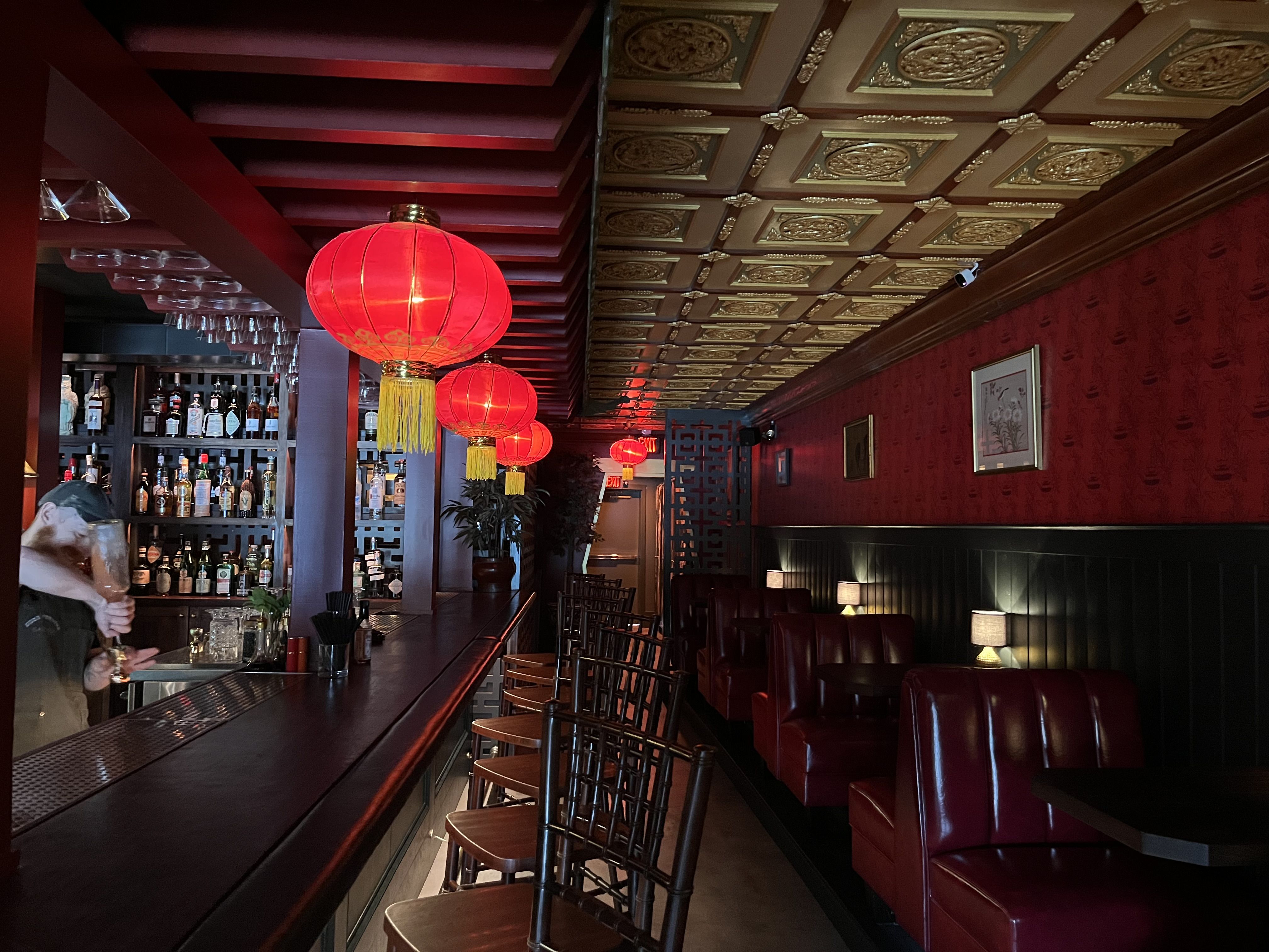 Dim bar with a long counter, red Chinese lanterns, tasseled gold ceiling panels, red wallpaper, and liquor bottles on shelves behind the bar; red leather booths along the wall.