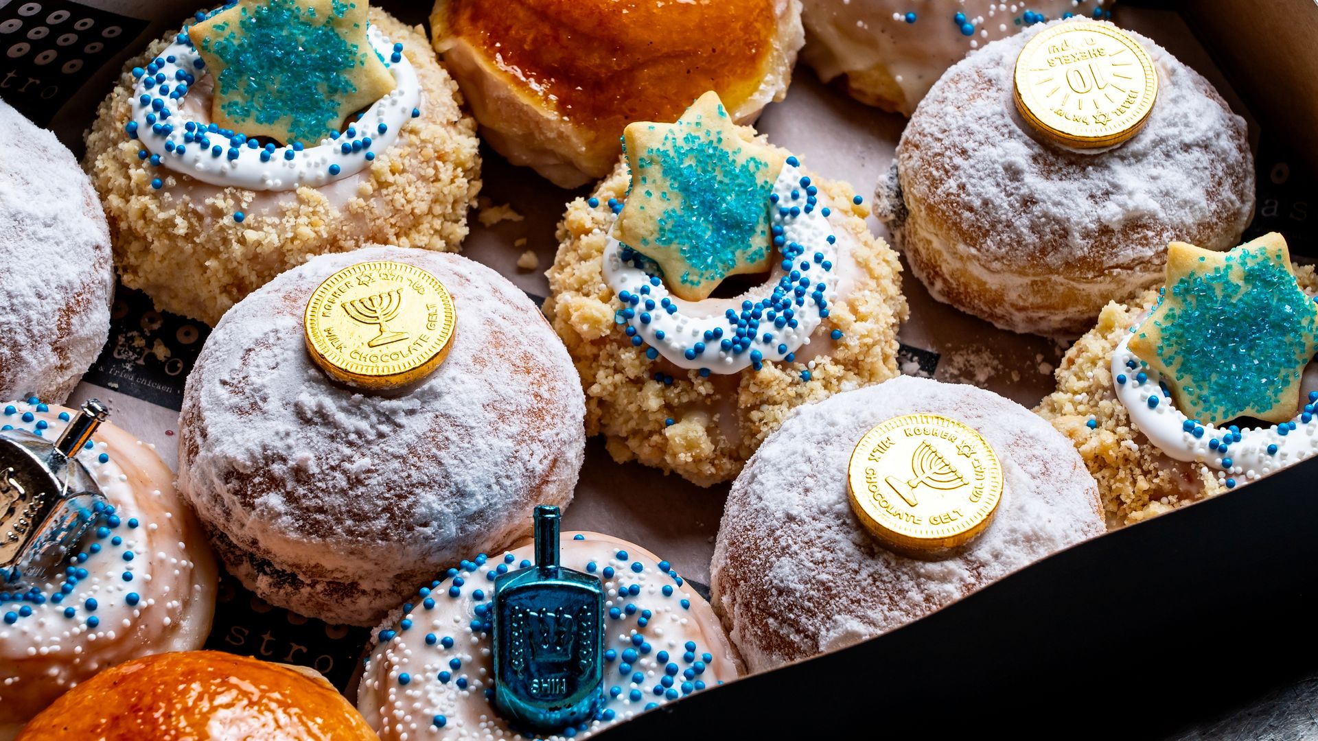 Box of assorted Hanukkah donuts decorated with powdered sugar, blue and white sprinkles, gelt coins, chocolate dreidels, and star-shaped cookies with blue sugar crystals.