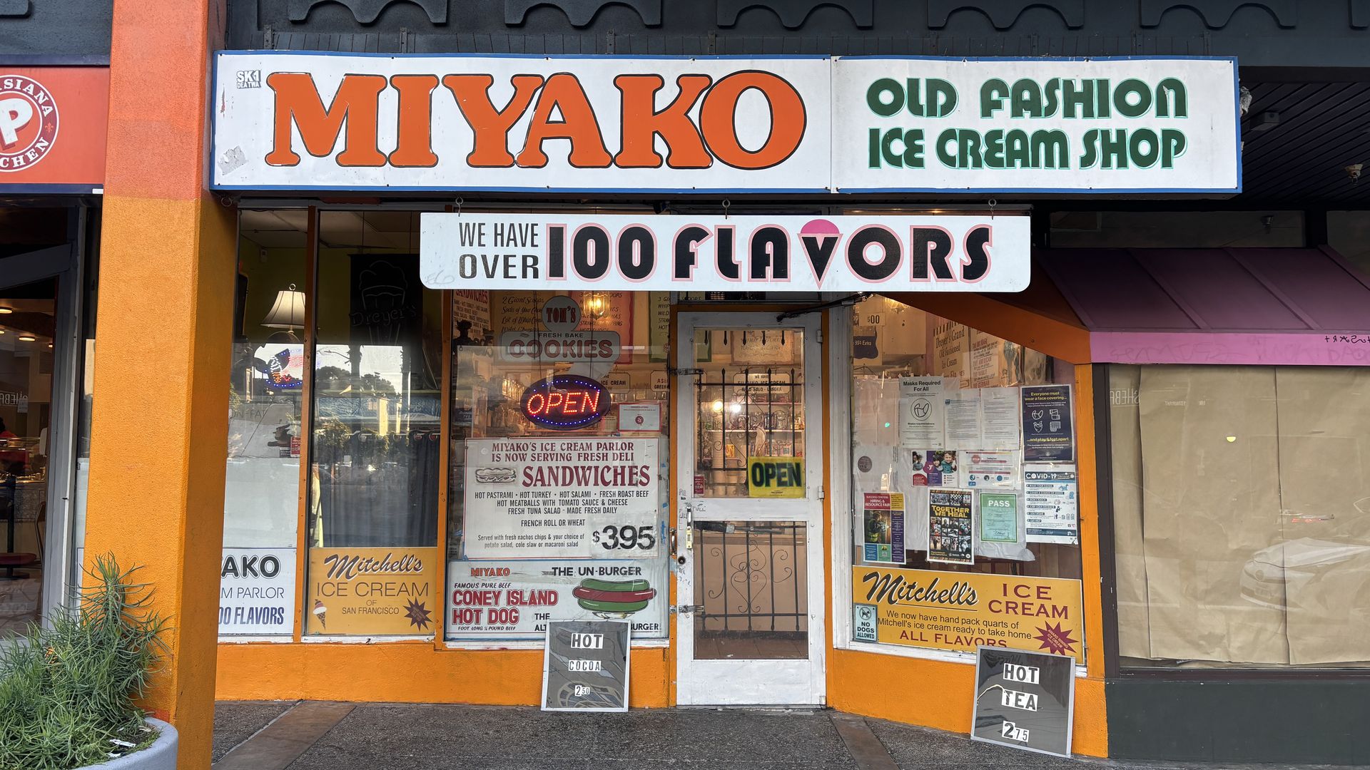 Front of Miyako Old Fashion Ice Cream Shop with orange trim and signs advertising over 100 ice cream flavors, sandwiches, Coney Island hot dog, Mitchell's Ice Cream, and hot cocoa and tea.