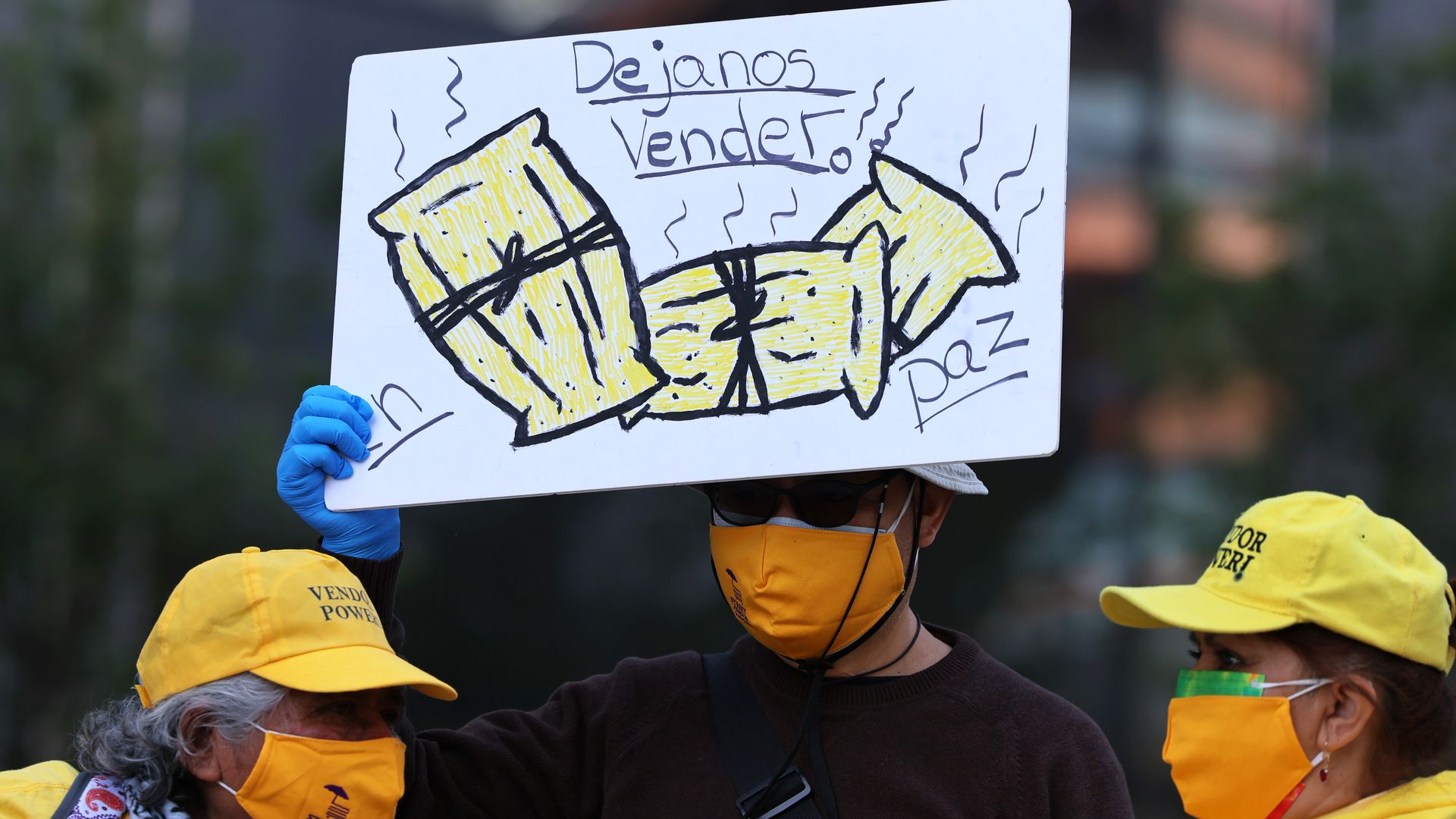 A man wearing a dark yellow mask holds up a sign with pictures of tamales. The sign is in Spanish and says "let us work" and "peace." Two women also wearing masks stand to both of his sides.