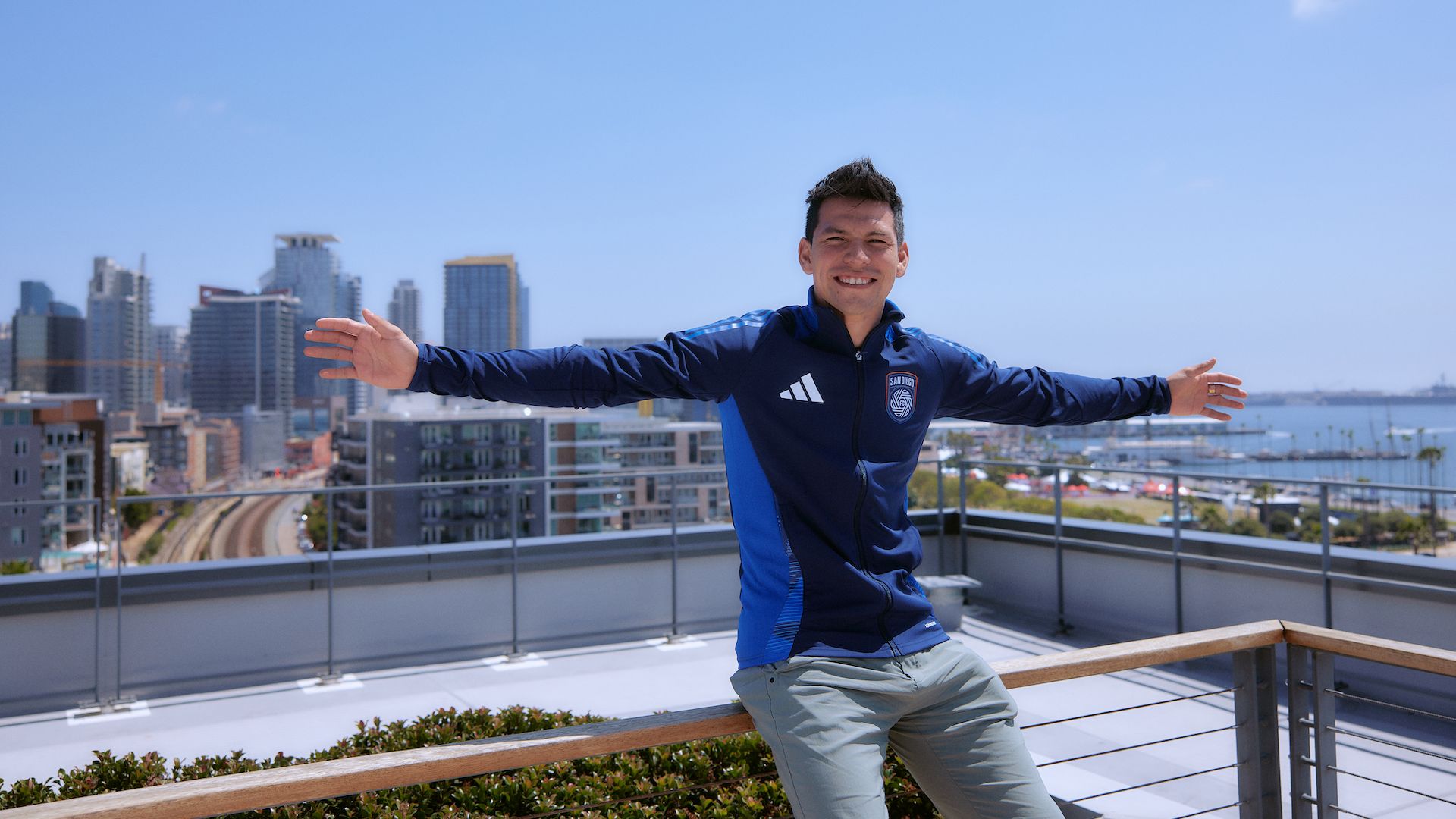 A soccer player leans on a rooftop railing with his arms stretched out with the city skyline and San Diego Bay behind him. 