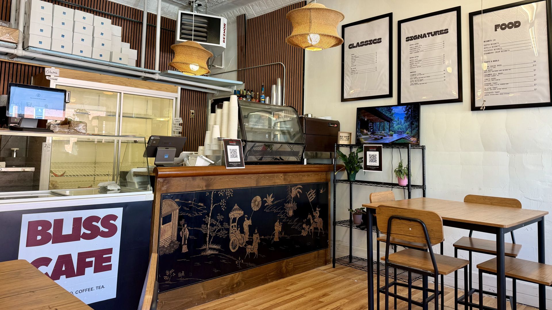 Cafe interior with wooden wall panels, white boxed storage on the left, two wicker pendant lights, a glass display counter, and framed menus reading "CLASSICS", "SIGNATURES", and "FOOD" on the right.