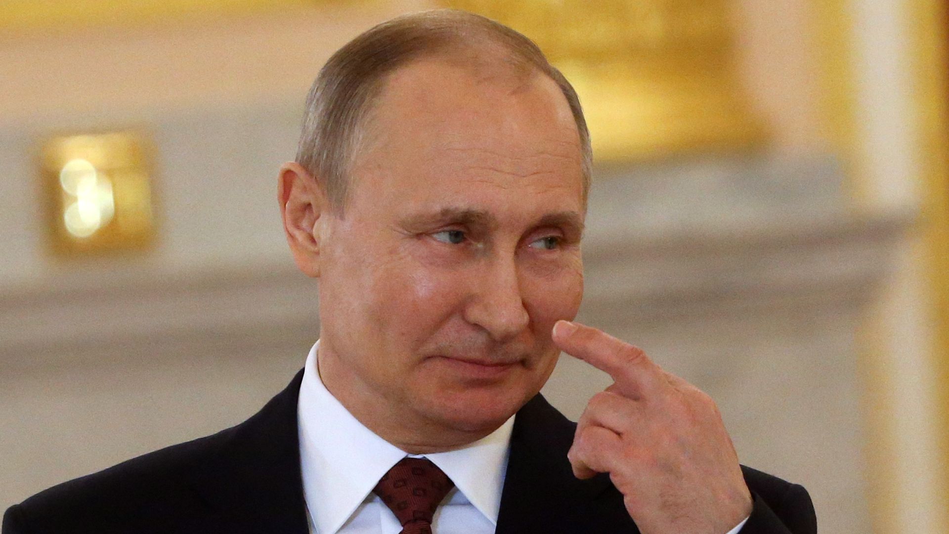 Vladimir Putin in a suit smiles to someone in the distance and looks sideways out of his eyes and points to himself, before a gold background.