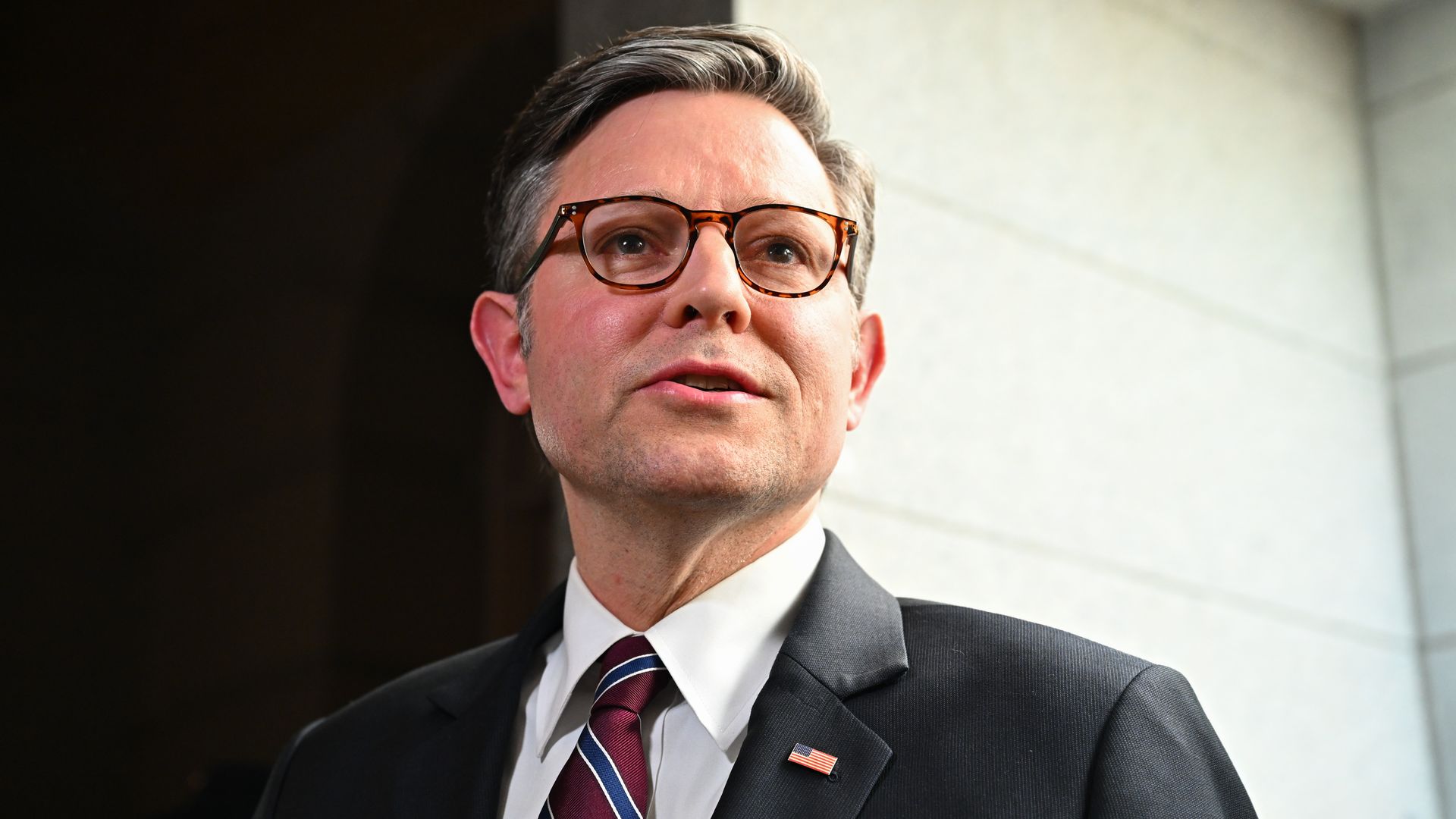 Close-up of a man in a dark suit and striped burgundy tie, wearing tortoiseshell glasses. He looks upward, with a light stone wall behind him and an American flag lapel pin.