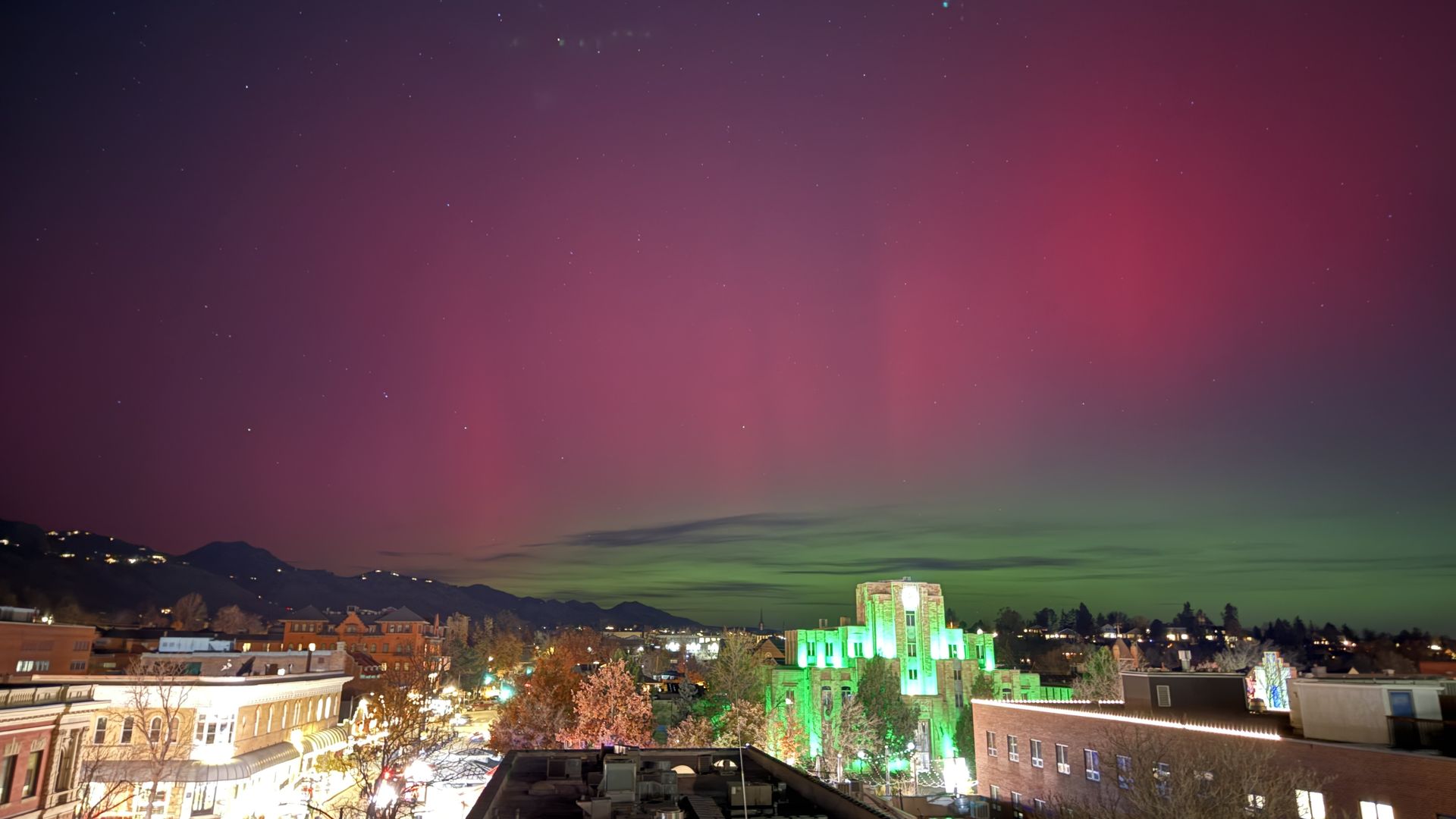 Night sky over a city with visible stars and a vibrant aurora showing green and pink colors above buildings and mountains in the background.
