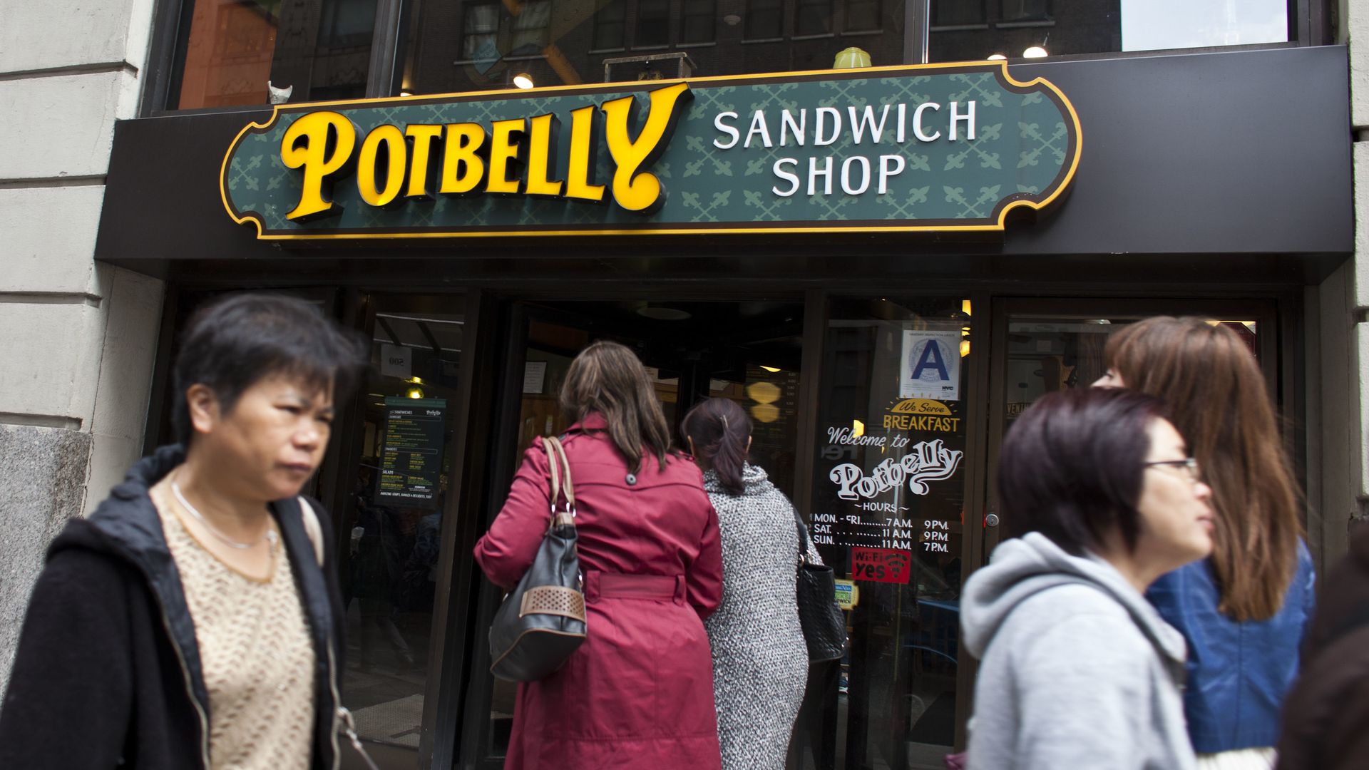 A Potbelly sign with the name in yellow and "sandwich shop" in white with people walking past the storefront.