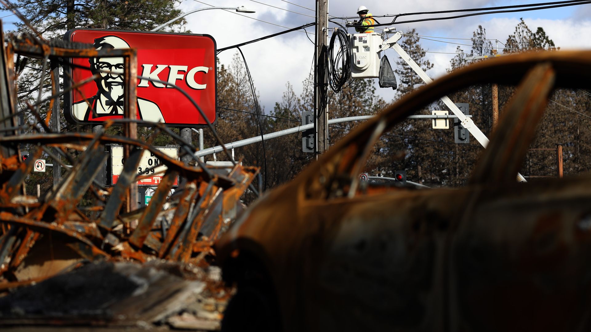 In this image, burned and wrecked cars sit in the foreground of the picture while in the background, a worker tries to fix electricity on a telephone pole.
