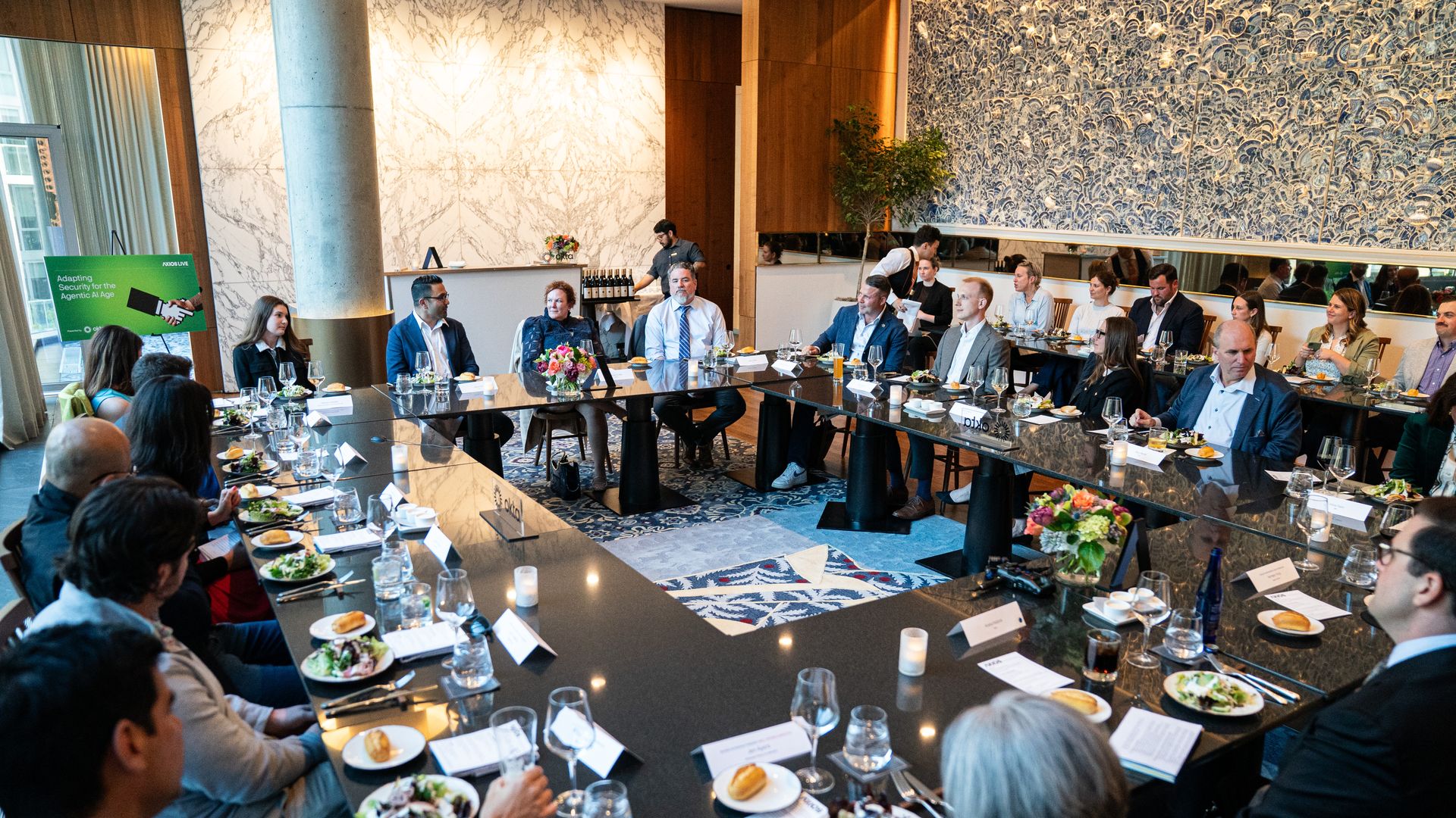 Business luncheon in a stylish conference room; professionals in suits sit around a long glossy table with plates, wine, and name cards, marble walls, and a server in the background.