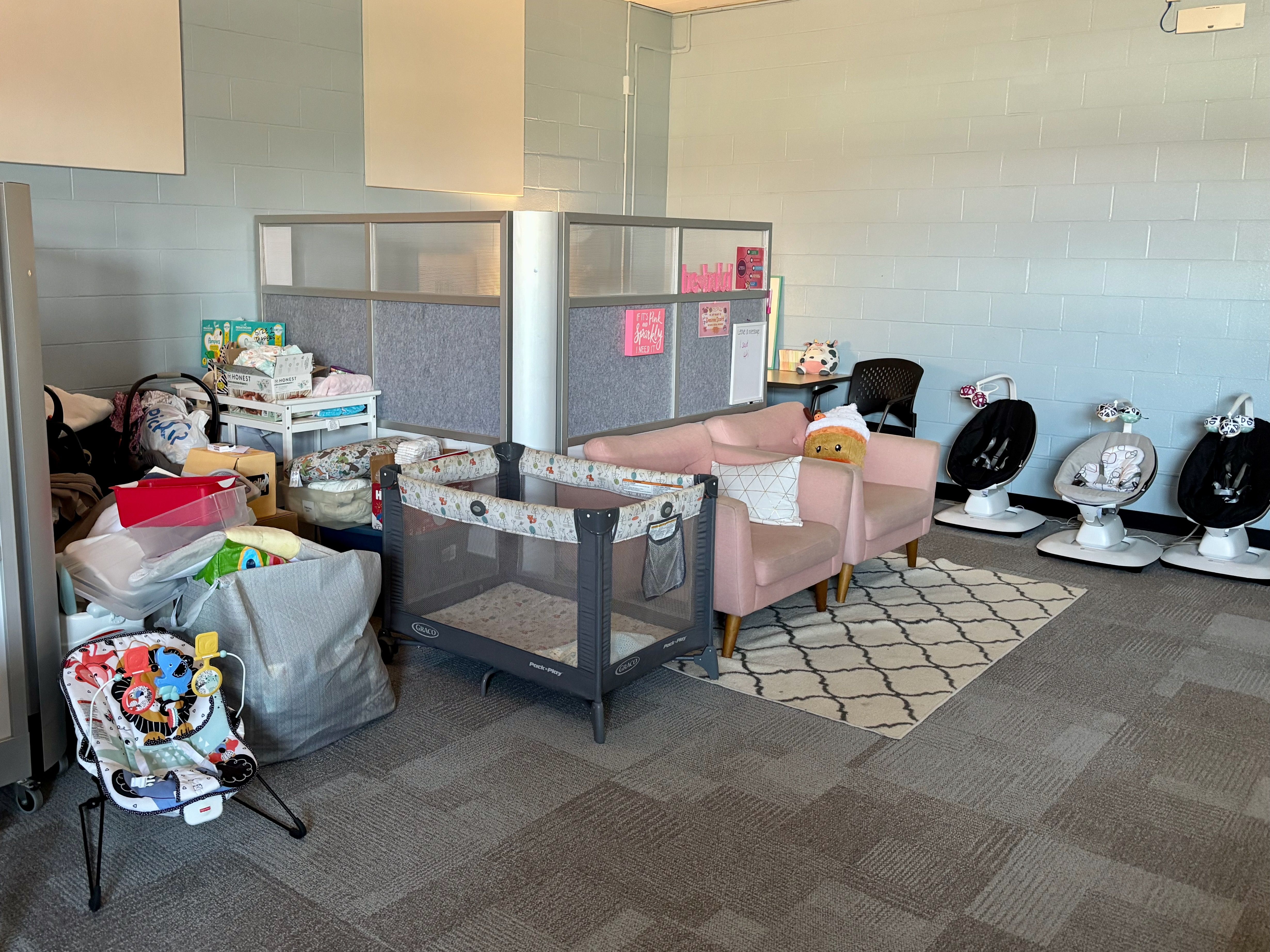 Bright room with light blue walls, cluttered baby area. A gray playpen sits by a pink loveseat, a geometric rug, and three white infant chairs along the wall; left side filled with bags and boxes.