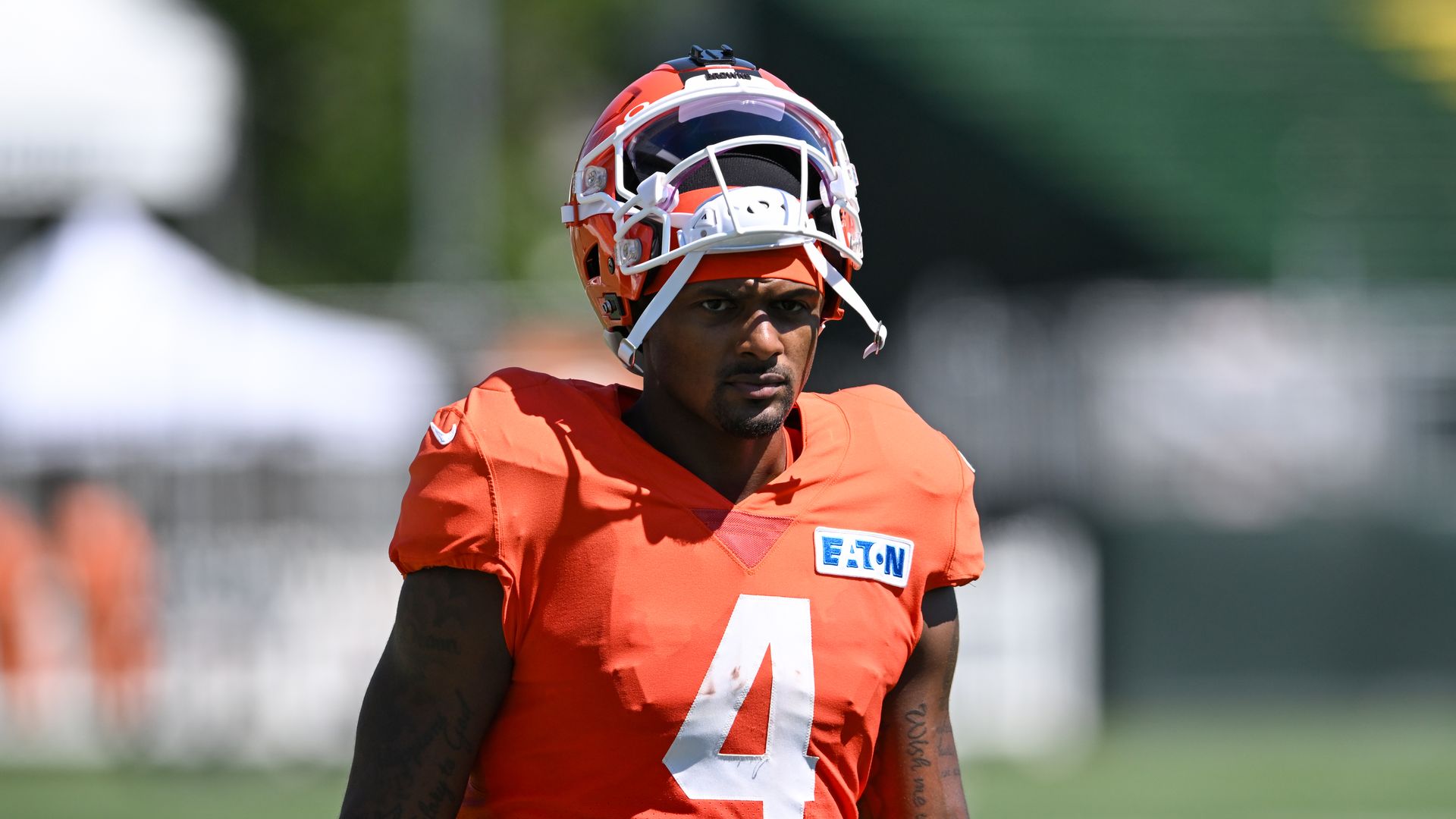Male football player in an orange jersey with the number 4 and a white-and-orange helmet, standing on a sunny practice field.
