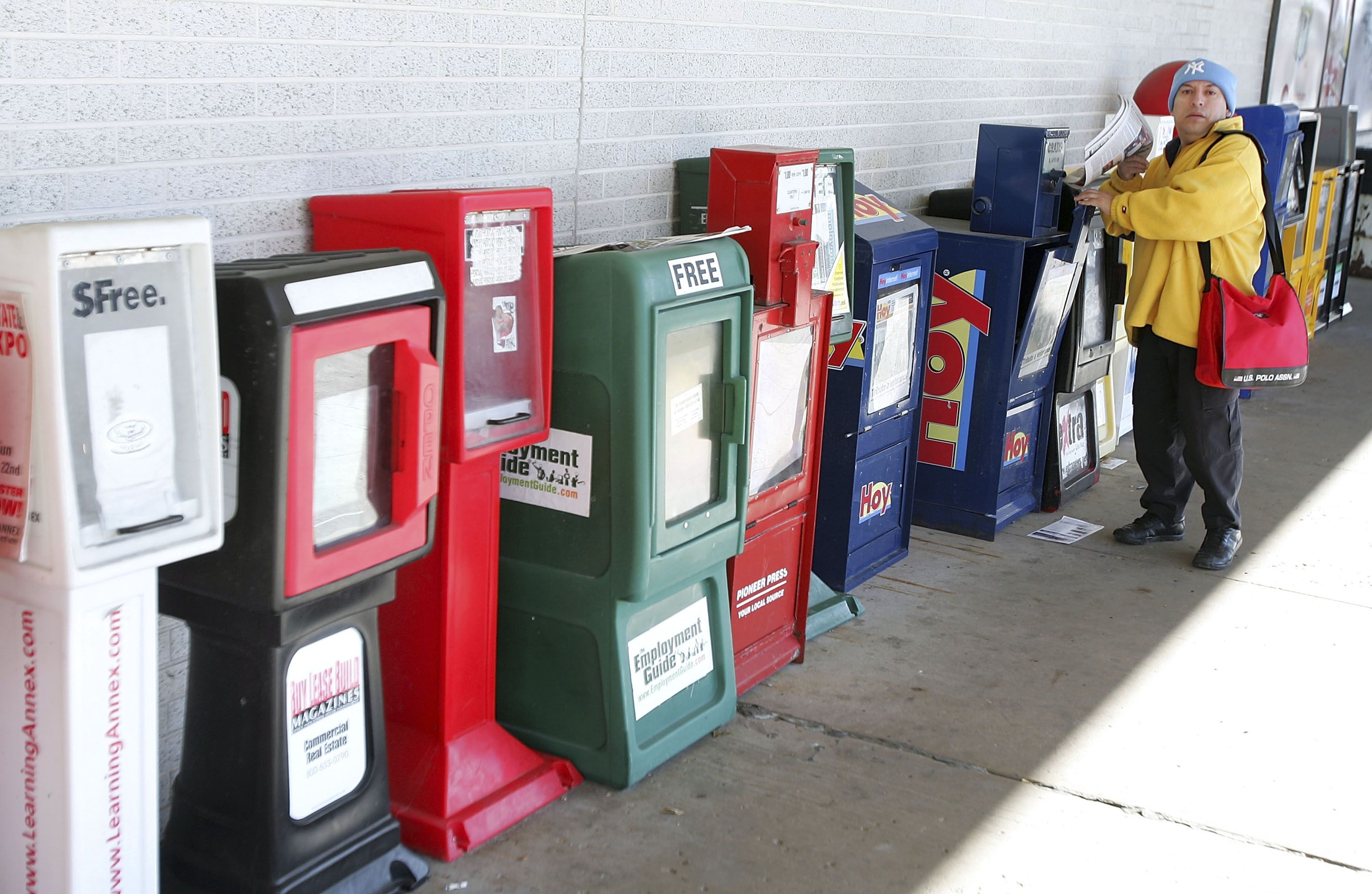 Man in yellow hoodie, blue beanie, and red bag picking up newspaper from a row of colorful newspaper vending machines outside against a white brick wall.
